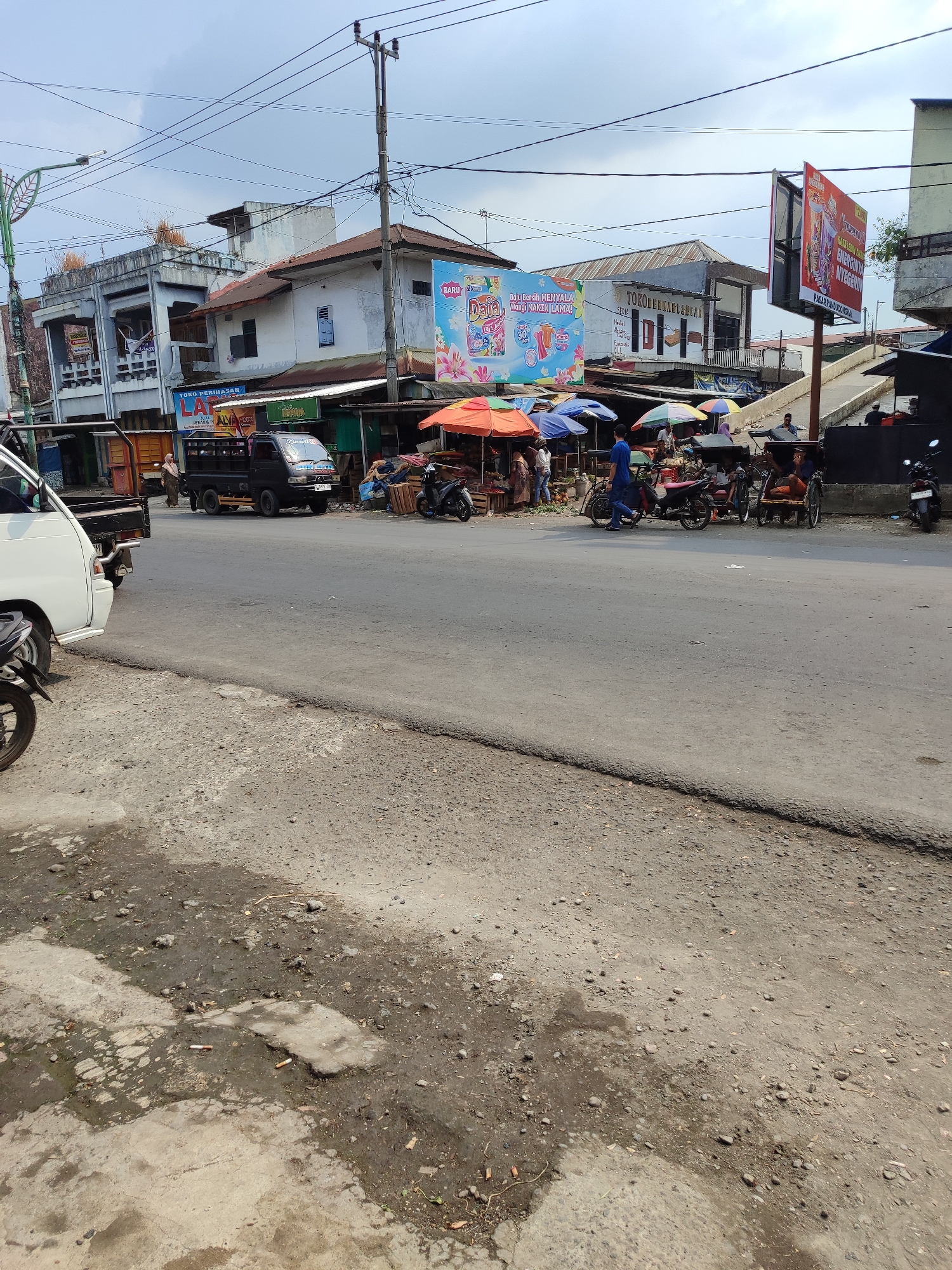 The image depicts a street scene in Randudongkal, Indonesia, characterized by a bustling market area alongside a paved road. In the foreground, a portion of a concrete surface with scattered debris and some damp patches is visible, suggesting an outdoor environment. Across the road, a market with numerous stalls covered by colorful umbrellas is in full swing. Various goods, likely produce and other merchandise, are displayed on tables and on the ground. Several individuals are present, some engaged in shopping or selling, while others are seen operating or standing near motorcycles and a three-wheeled vehicle.
Dominating the background are several buildings, ranging from multi-story structures with balconies and signs to simpler shops with corrugated metal roofs. Prominent advertising billboards are mounted on poles, advertising products and services. Power lines crisscross the sky above the scene. Several vehicles, including a white pickup truck and a black pickup truck, are parked or moving on the road. The sky is partly cloudy, indicating daytime. The overall impression is that of a typical commercial hub in a local Indonesian town.