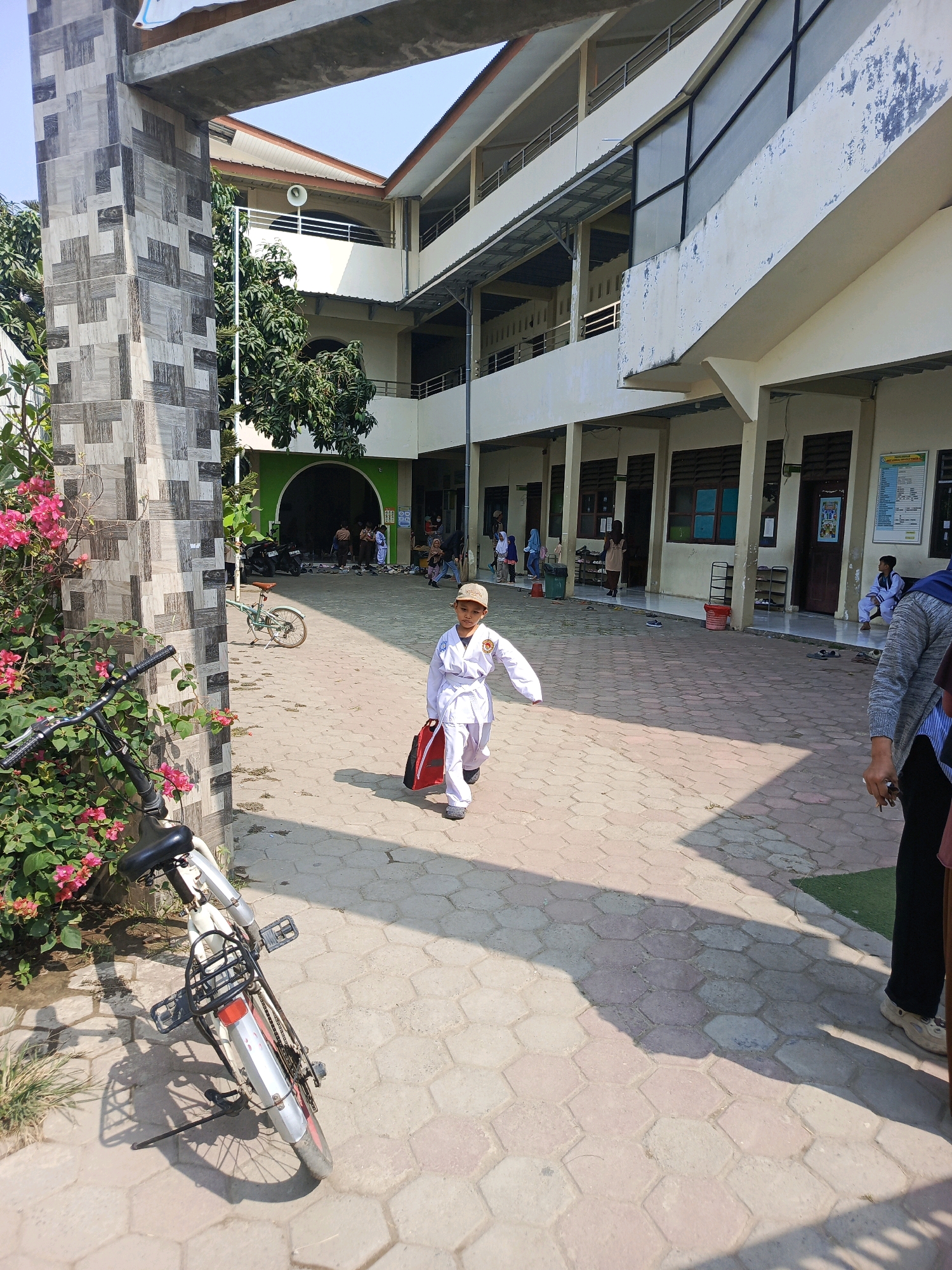 The image depicts an outdoor courtyard at what appears to be a school in Jati Mulya, Indonesia, likely a primary or secondary educational institution. A young child, dressed in a white karate uniform with a belt and a cap, is walking towards the camera, carrying a red bag. The child is in motion, suggesting they are either arriving or departing. In the foreground, to the left, a white bicycle is parked near a wall adorned with patterned tiles and flowering bushes with pink blossoms. The courtyard is paved with hexagonal grey and tan interlocking bricks. In the background, a multi-story building with white walls and balconies is visible. Several other individuals, including children and adults, are present in the background, some near doorways and others standing or sitting. The environment suggests a bright, sunny day with clear blue skies. The architecture and general atmosphere are consistent with a public school or community facility in a Southeast Asian urban setting.