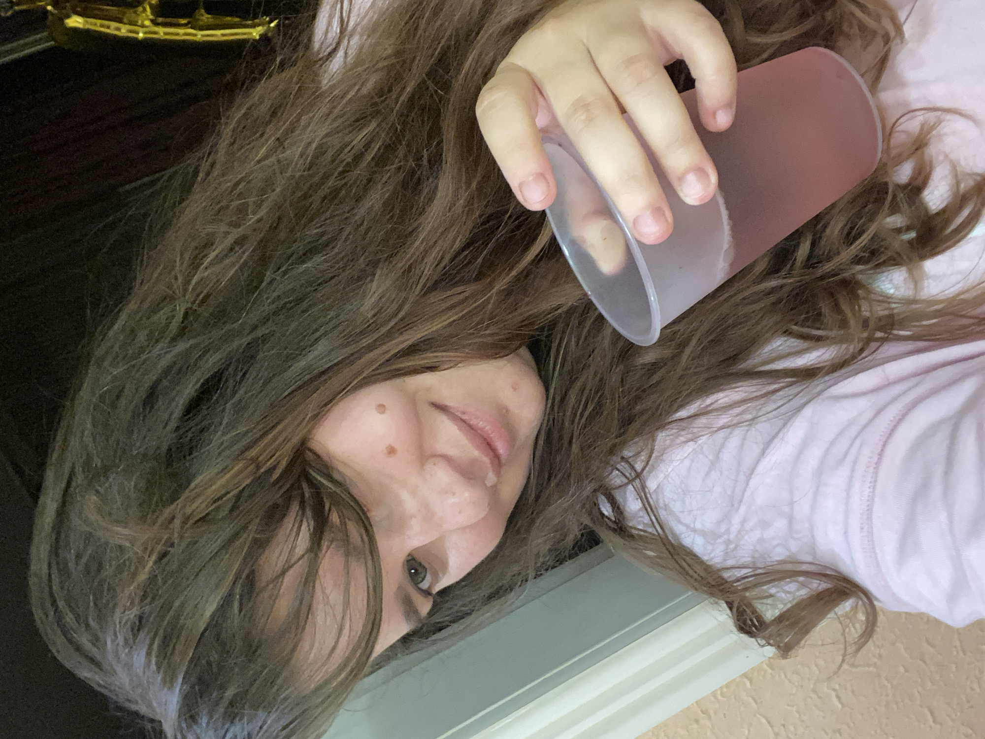 A medium close-up selfie shows a young woman with long, wavy brown hair and fair skin holding a clear plastic cup of pink liquid in her right hand. She is wearing a light pink t-shirt. Her face is tilted, and she is looking at the camera. She has freckles and two prominent moles on her right cheek. The background consists of a textured, light-colored wall on the left and a dark background with a glimpse of a gold decoration on the right. The scene appears to be indoors, possibly in a casual setting. No specific details about the city of Perm, Russia are visible in the image.