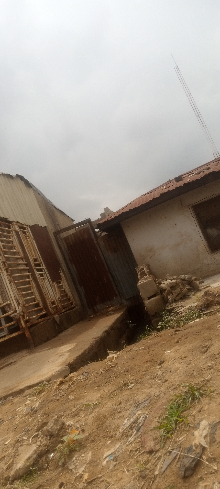 The image shows a deteriorated street scene in Sabon Tasha, Nigeria. Two buildings with corrugated iron roofs are visible. One has rusty metal doors and window coverings; the other has a window and broken concrete supports. A dirt ground dominates the foreground and contains debris. An open trench runs between the buildings and the foreground. The sky is overcast. A communication tower is barely visible in the upper right.