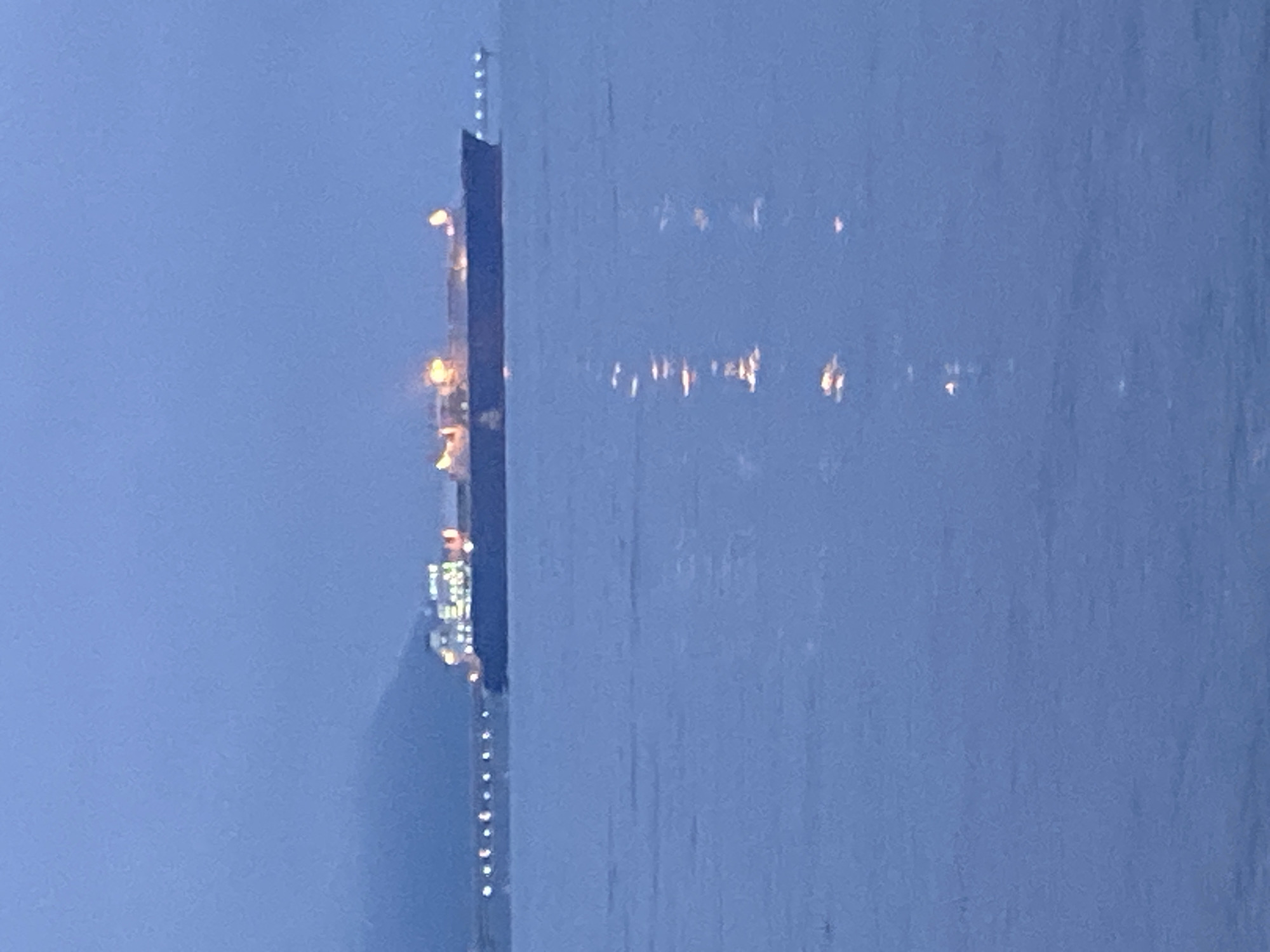 The image shows a large ship, likely a cargo vessel or tanker, at sea near the city of Korsakov, Russia. The ship is illuminated by artificial lights, suggesting it is either docked or navigating in the early morning or evening. The water is calm, with reflections of the ship's lights visible on the surface. In the background, a dimly lit shoreline or landmass can be seen, possibly a portion of Korsakov itself. The overall atmosphere is serene and somewhat muted due to the low light conditions.