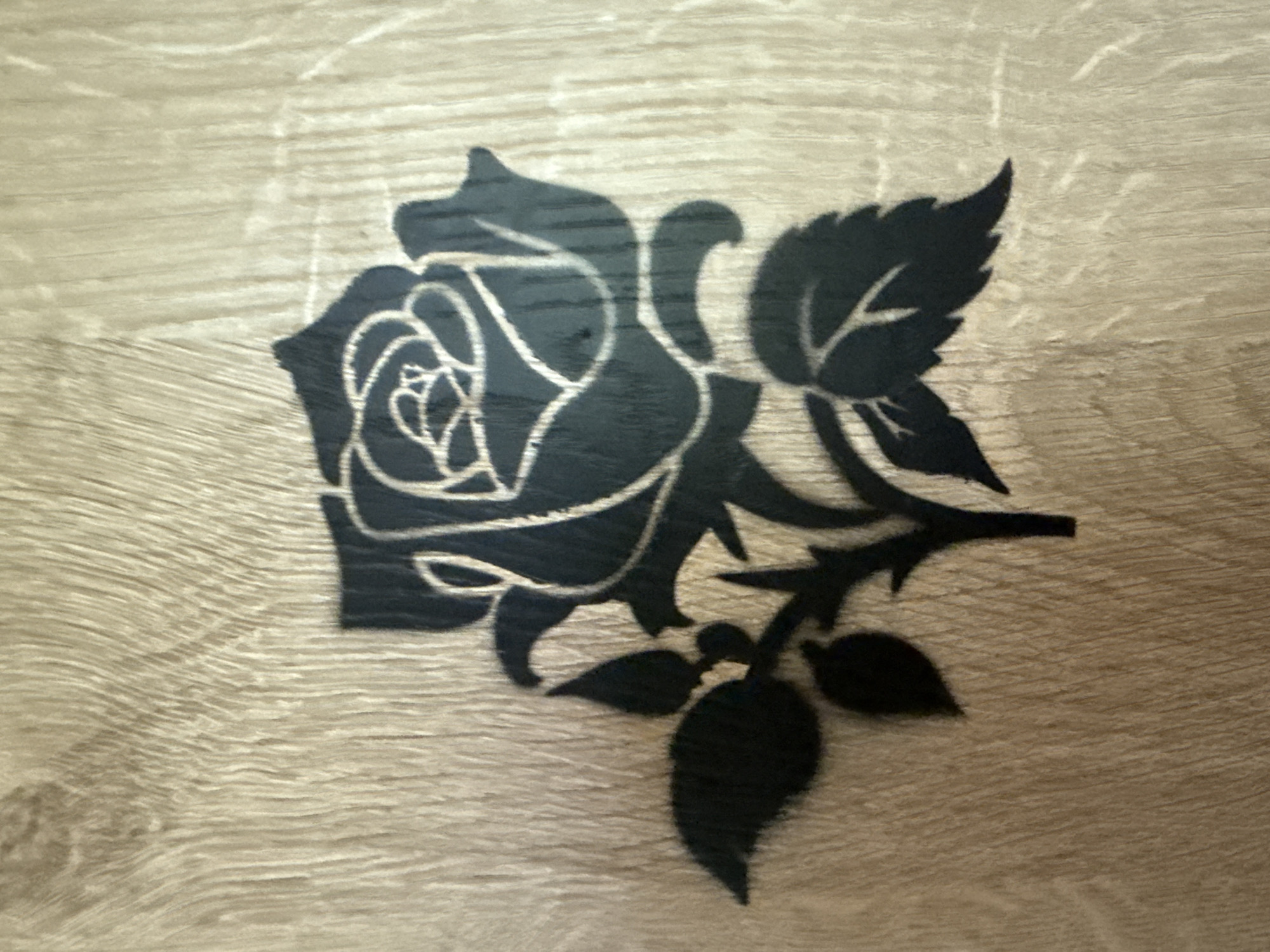 The image displays a stencil of a dark rose with white outlines against a light, textured wooden surface. The rose is depicted with its petals unfurled and a stem with leaves and a bud. The background shows a wood grain pattern with vertical lines, suggesting a close-up view of a wooden surface such as a table or wall. There are no people or animals present in the image. The scene is static and does not depict any actions or interactions. The location is not evident from the image itself, and therefore, cannot be described as being in Galinė, Lithuania.