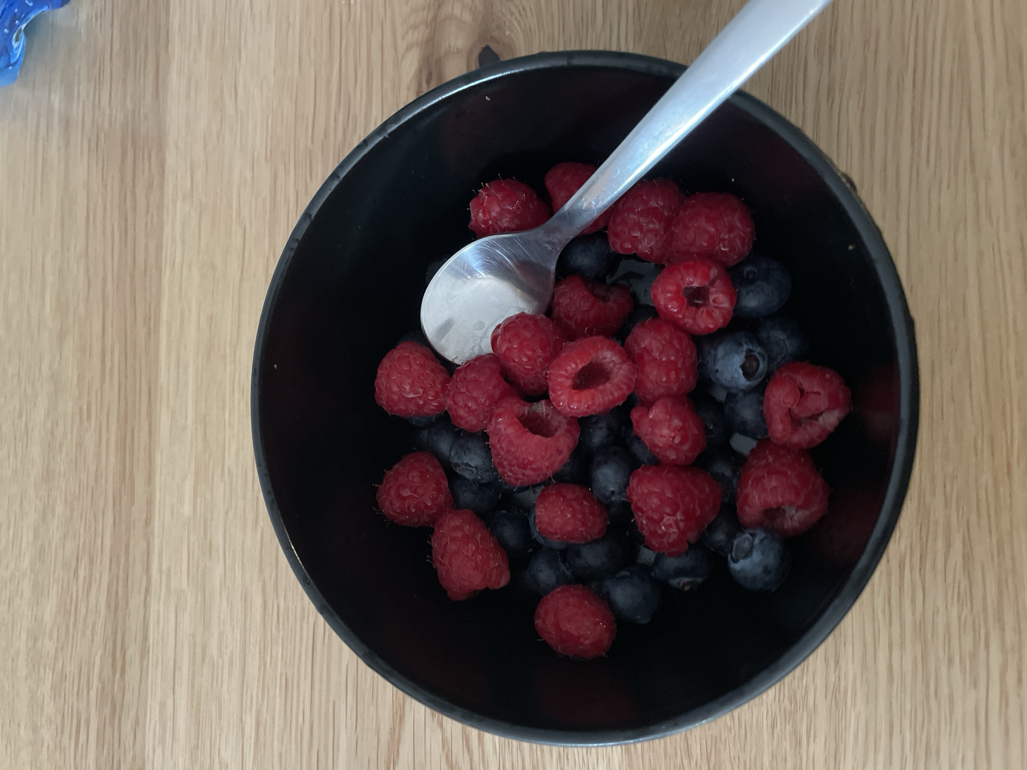A black bowl containing raspberries and blueberries sits on a wooden surface. A silver spoon rests inside the bowl. The scene's location within the city of Sofia, Bulgaria is indeterminable from the image.