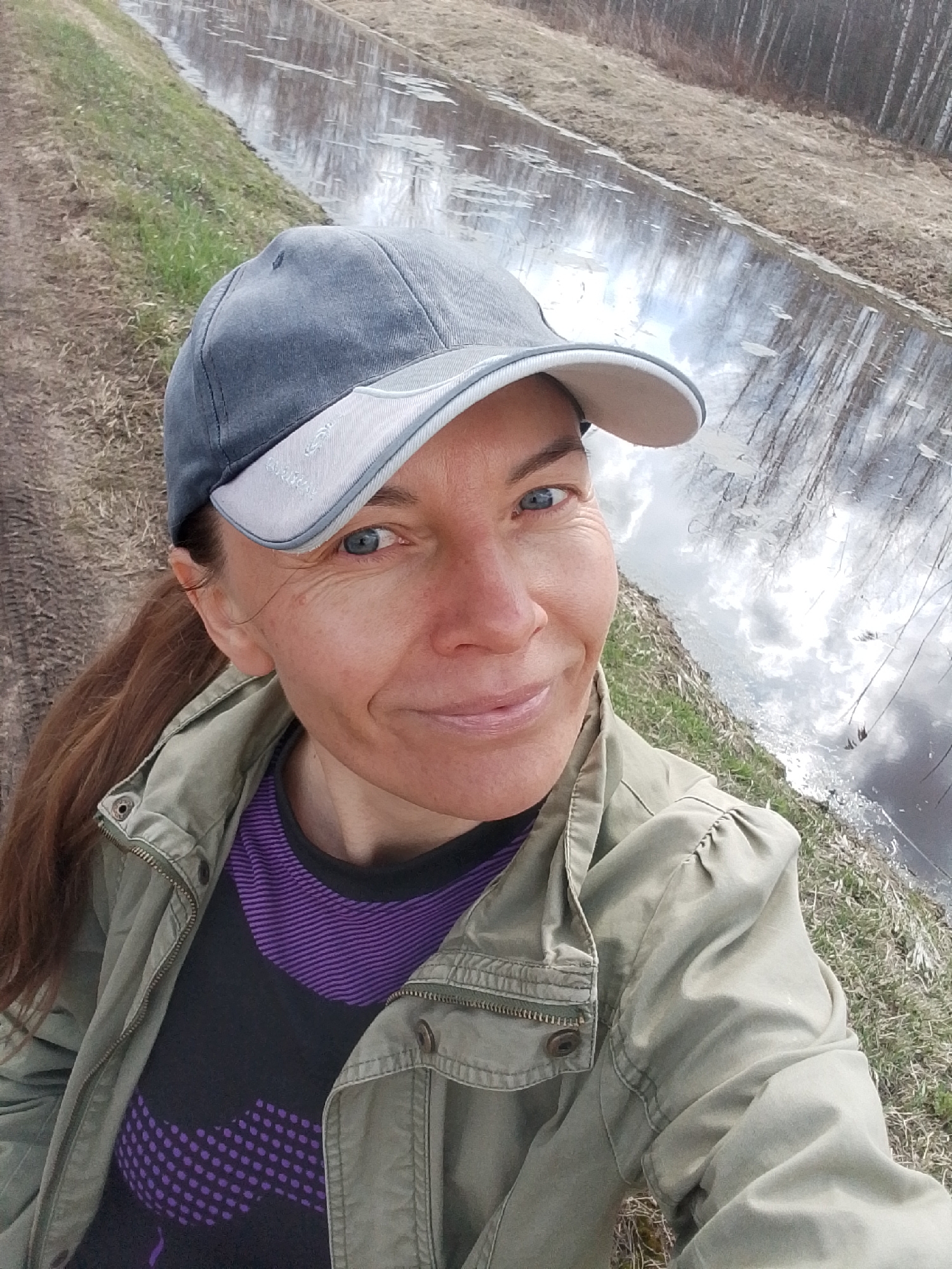 A woman with blue eyes and brown hair pulled into a low ponytail is taking a selfie. She is wearing a grey and light grey baseball cap, a black t-shirt with purple dots and stripes, and a green jacket. Behind her is a long, narrow ditch or canal filled with water that reflects the sky and trees. The banks of the ditch are covered with dry grass and brush. In the far background are trees without leaves. The location is Снопок Новый, Russia.