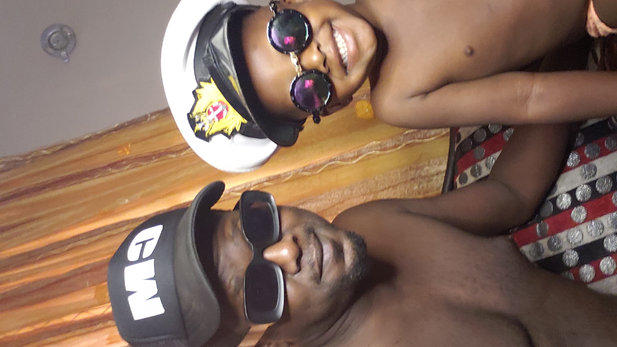 This medium close-up image, oriented sideways, captures an adult male and a child posing for a photograph while lying down in Sabon Tasha, Nigeria. Both individuals have dark skin tones and are shirtless.
The adult, closer to the viewer, wears a black baseball cap with white letters, possibly "EW" or "CW," and large, rectangular black sunglasses. He has a beard or stubble.
Behind him, a child, smiling broadly, wears a white captain's hat with a black brim and a gold emblem featuring a red design. The child also sports round, reflective black sunglasses.
They are resting on patterned surfaces; beneath the adult is a fabric with red, white, and black stripes interspersed with silver circular motifs, while the area behind the child shows a plain orange-brown fabric. A light-colored ceiling with a recessed light fixture is visible in the upper part of the background. The child's visible smile conveys a joyful and relaxed mood.