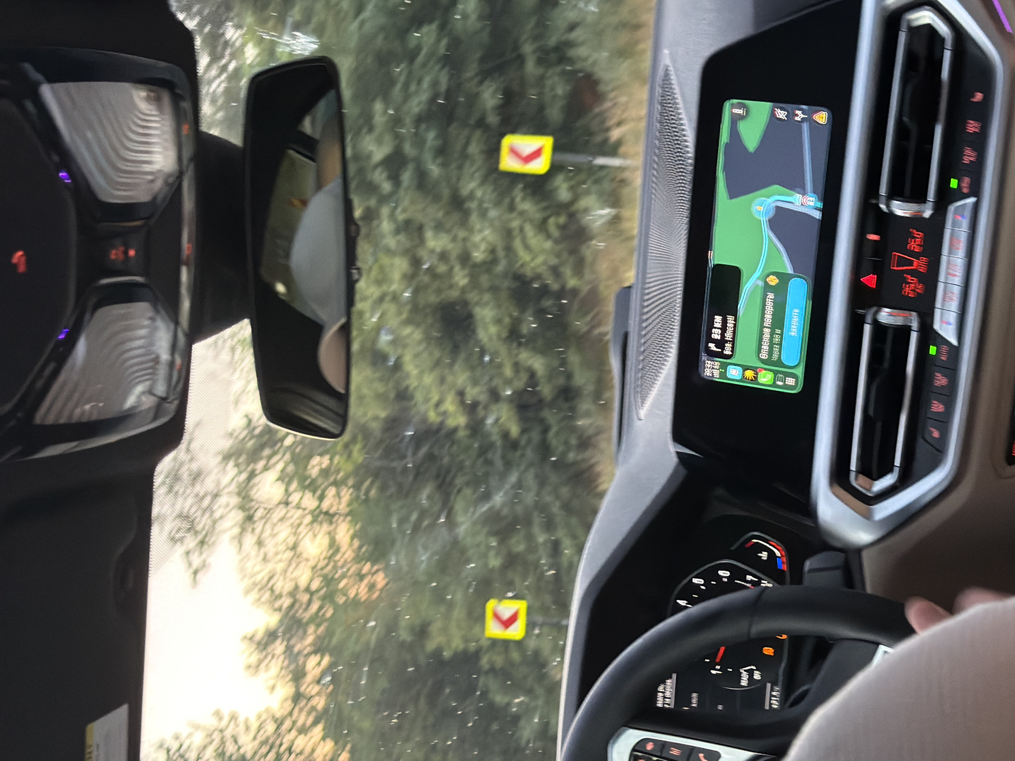 The image depicts the interior of a car, looking out through the windshield. The driver's hand is visible on the steering wheel. The car's dashboard displays navigation information on a screen, showing a map with a route highlighted and indicating "23 KM" to "Şaşca, Hîncești," and a warning about "Dangerous turns." There are also climate control buttons and displays visible. Outside the car, trees are visible, suggesting a rural or wooded area. A yellow directional sign with a black arrow pointing left is mounted on a pole and visible through the windshield. The sky in the background is bright, possibly indicating dawn or dusk. The context suggests the car is driving on a road near Fundul Galbenei, Moldova, as indicated by the navigation display.