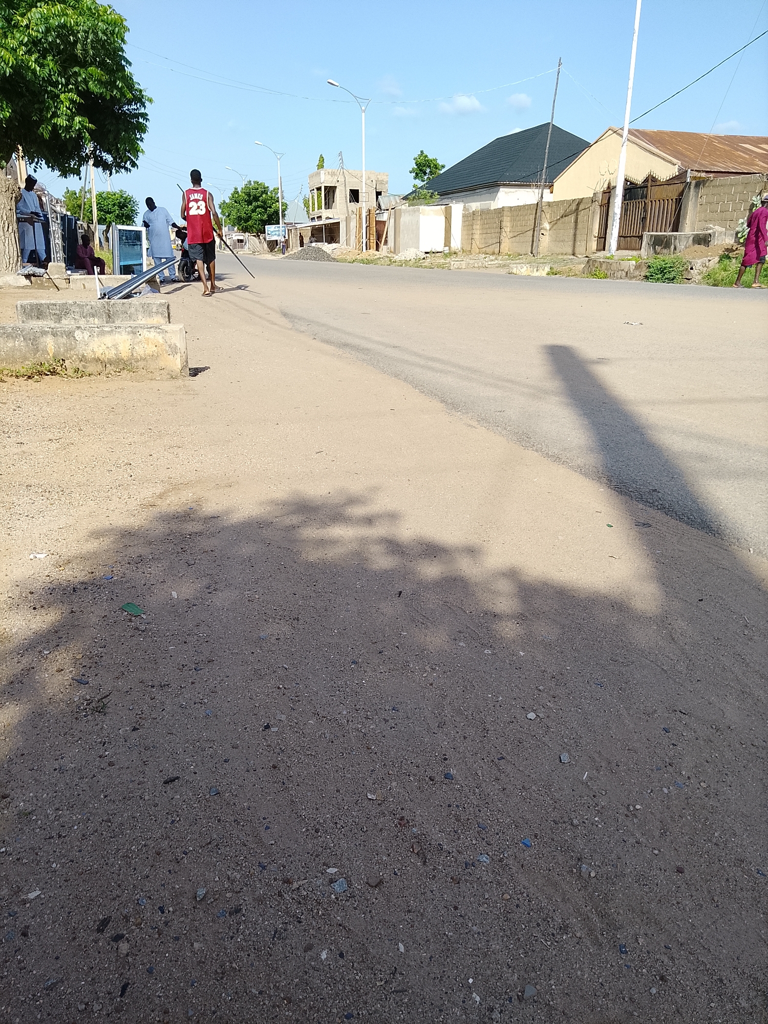 A low-angle, full shot captures a street scene in Wauro Leggal, Nigeria. In the foreground, the sandy, gravelly texture of the road dominates, with a patch of concrete curb to the left and distinct shadows cast by trees and a utility pole, indicating a sunny day. The midground features a paved road with a distinct shadow of a pole stretching across it. On the left side of the road, near the curb, several individuals are gathered around what appears to be merchandise, possibly appliances or display units, with one person in a red basketball jersey with "JAMES 23" visible. A motorcycle is parked behind them. Beyond the people, construction is evident on the right side of the road, with partially built structures and materials. The background reveals residential buildings with varying roof types and brick walls, under a clear blue sky. The overall impression is that of a bustling, albeit partially under construction, urban environment.