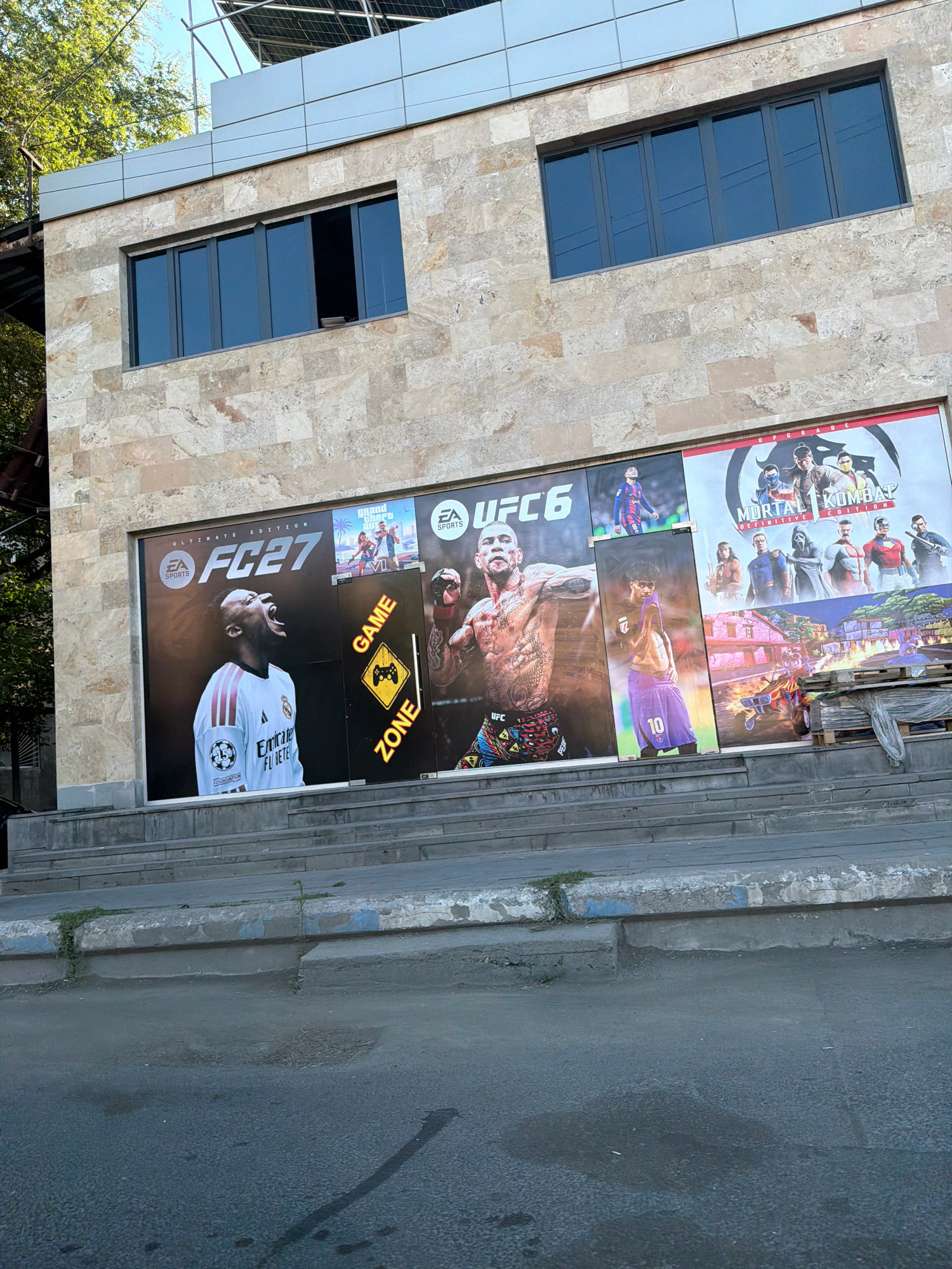 The image shows a building in Yerevan, Armenia, with several large posters advertising video games displayed on its facade. The building has a stone exterior and several windows. The posters feature images from games such as FC 27, UFC 6, and Mortal Kombat. One poster depicts a person in a white sports jersey with the text "Emirates FL BE". Another poster shows a muscular fighter with tattoos. There is also a sign with a yellow triangle and a joystick symbol, labeled "GAME ZONE". In the foreground, there are steps leading up to the building and a street with some wear and tear. The overall scene suggests a place where video games are sold or promoted, possibly a store or entertainment center.