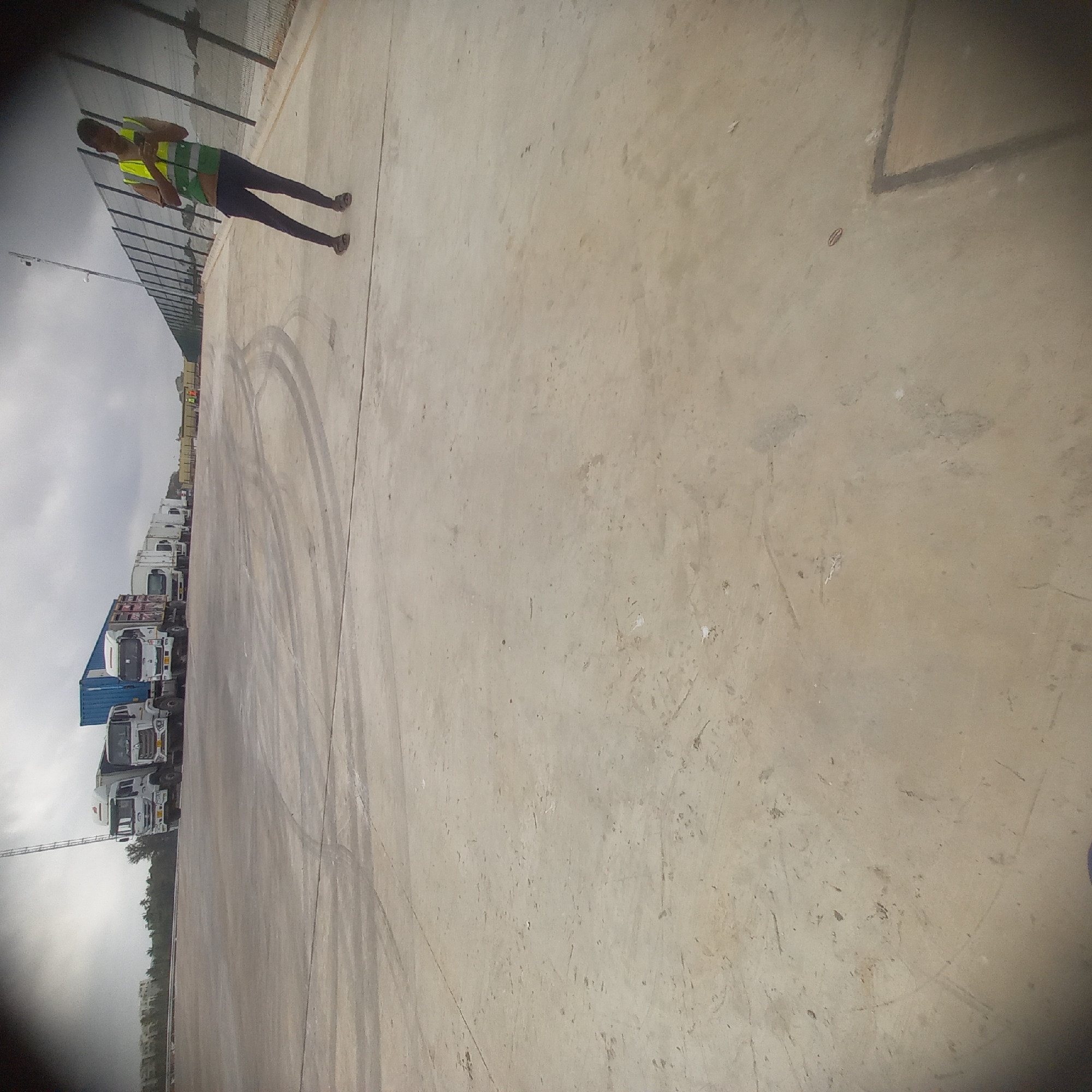 The image depicts a paved outdoor area, possibly a port or industrial zone, in Bububu, Tanzania. A line of white and blue container trucks is parked on the left side of the frame. In the mid-ground, on the right, a person wearing a yellow and green high-visibility vest and dark pants is standing near a fence, looking at their phone. The ground is concrete with tire marks visible. The sky is overcast.