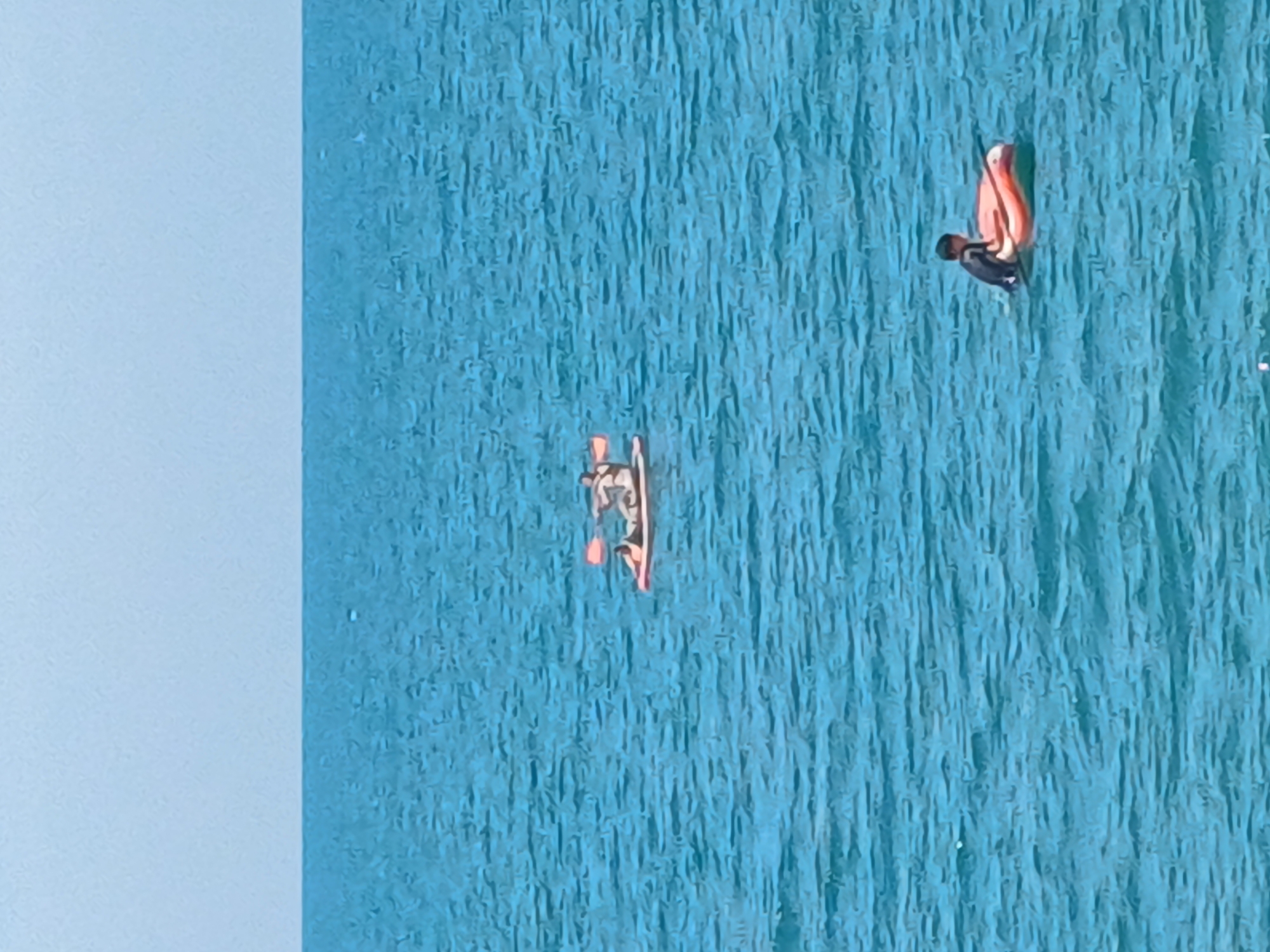 In Sochi, Russia, on a clear day, two people are enjoying the blue sea. In the foreground, a person wearing a black wetsuit sits in an orange inflatable tube. Further in the distance, two individuals are on a red and white paddleboard. One person is paddling with a red paddle, while the other is lying down on the board. The calm, rippling water stretches to the horizon, meeting a pale blue sky.