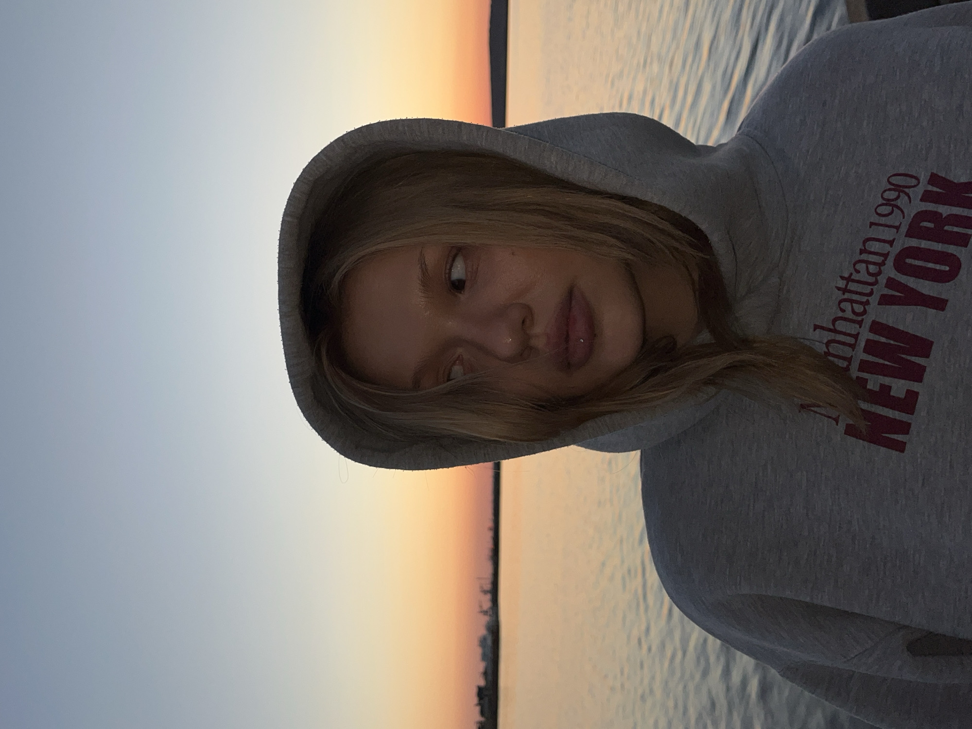 A woman wearing a grey hooded sweatshirt with the words "Manhattan 1990 NEW YORK" in red lettering stands outside at sunset, likely by the water, in Chaikovsky, Russia. The woman is facing to the right with her head tilted. The background shows a body of water and a sunset sky.