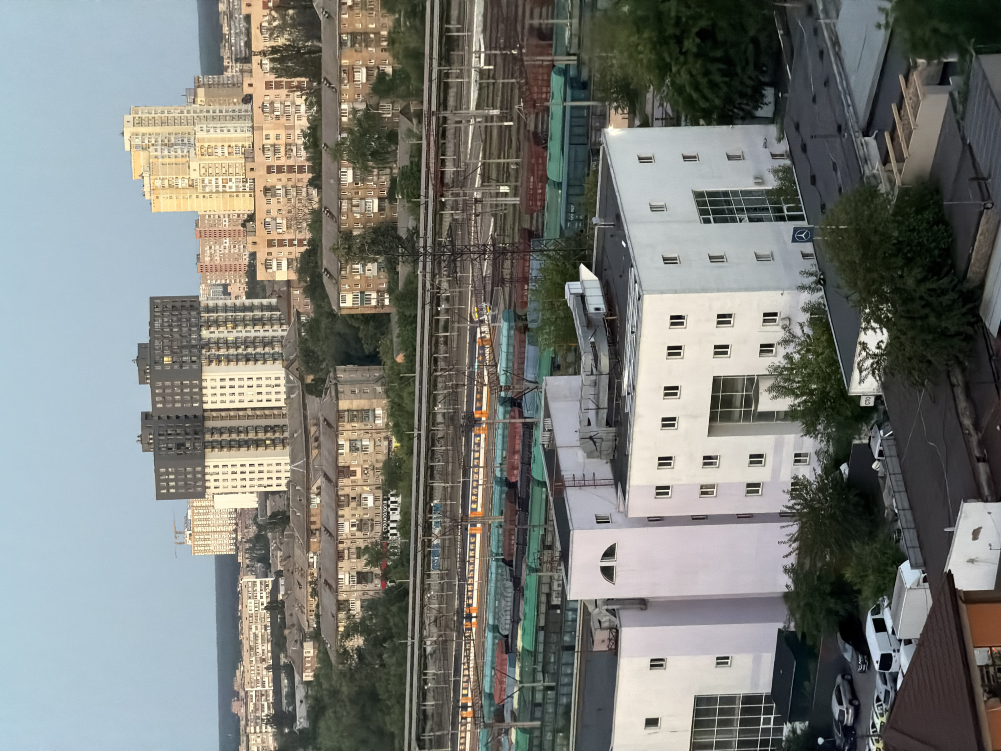 The image depicts an aerial view of a cityscape in Kyiv, Ukraine, likely taken from a high vantage point. In the foreground, several modern, white-facade buildings are visible, some with large glass windows. Adjacent to these buildings, a parking area contains a variety of vehicles, including cars and vans. Beyond the immediate foreground, a railway yard is prominent, with multiple train tracks and numerous freight cars of varying colors, including green, blue, and red, loaded with goods. A passenger train with an orange and white color scheme is also present on the tracks. Further in the background, a dense residential area with apartment blocks and older Soviet-era buildings stretches across the landscape. Several taller, contemporary apartment buildings stand out against the sky. The environment appears to be an urban area with a mix of residential and industrial elements, with a clear sky and lush green trees interspersed throughout.