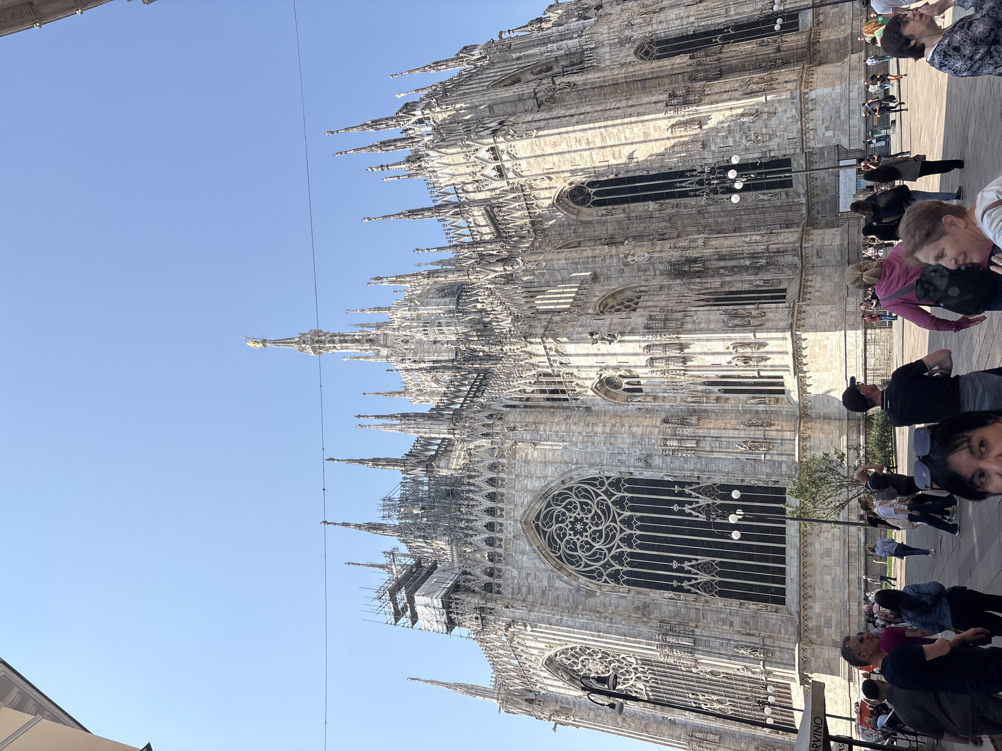The image shows the Milan Cathedral (Duomo di Milano) in Italy. The cathedral is a large, white stone building with intricate gothic architecture, featuring numerous spires and statues. Part of the building is under construction with scaffolding visible. A group of people are walking and standing on the sidewalk in front of the cathedral. The sky is blue.