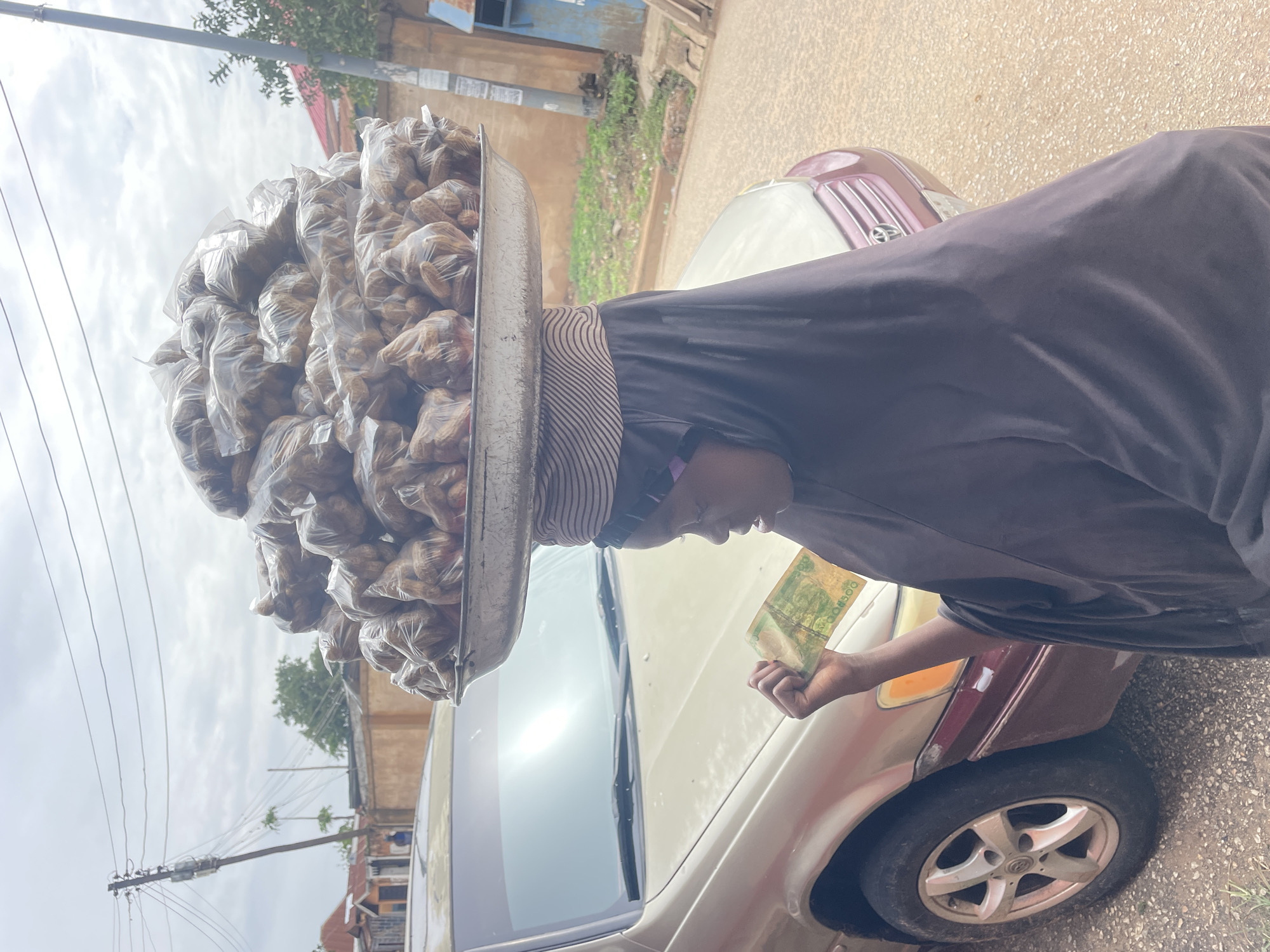 A street scene in Jos, Nigeria, depicts a daytime transaction under a partly cloudy sky. The central figure is a street vendor, seen in profile, wearing a dark garment and a striped head covering. They skillfully balance a large, silver-colored metal tray on their head, filled with numerous small, transparent plastic bags. Each bag contains brown, oval-shaped items, likely groundnuts (peanuts), suggesting the vendor is selling these snacks.
The vendor is actively engaged in a sale, looking down at a customer's hand, which extends from the right holding a green Nigerian N500 banknote. The transaction occurs beside a light-colored sedan car, which fills the lower and middle right of the frame, featuring a distinct maroon rear fender and a visible wheel.
The background shows overhead power lines and a utility pole, with distant buildings and green foliage indicating an urban or semi-urban setting. The ground beneath the car is a rough paved surface. The image captures a common element of daily life and street commerce in Nigeria, showcasing the traditional method of hawking goods. The time of day appears to be bright, and the overall scene reflects a typical, bustling environment.