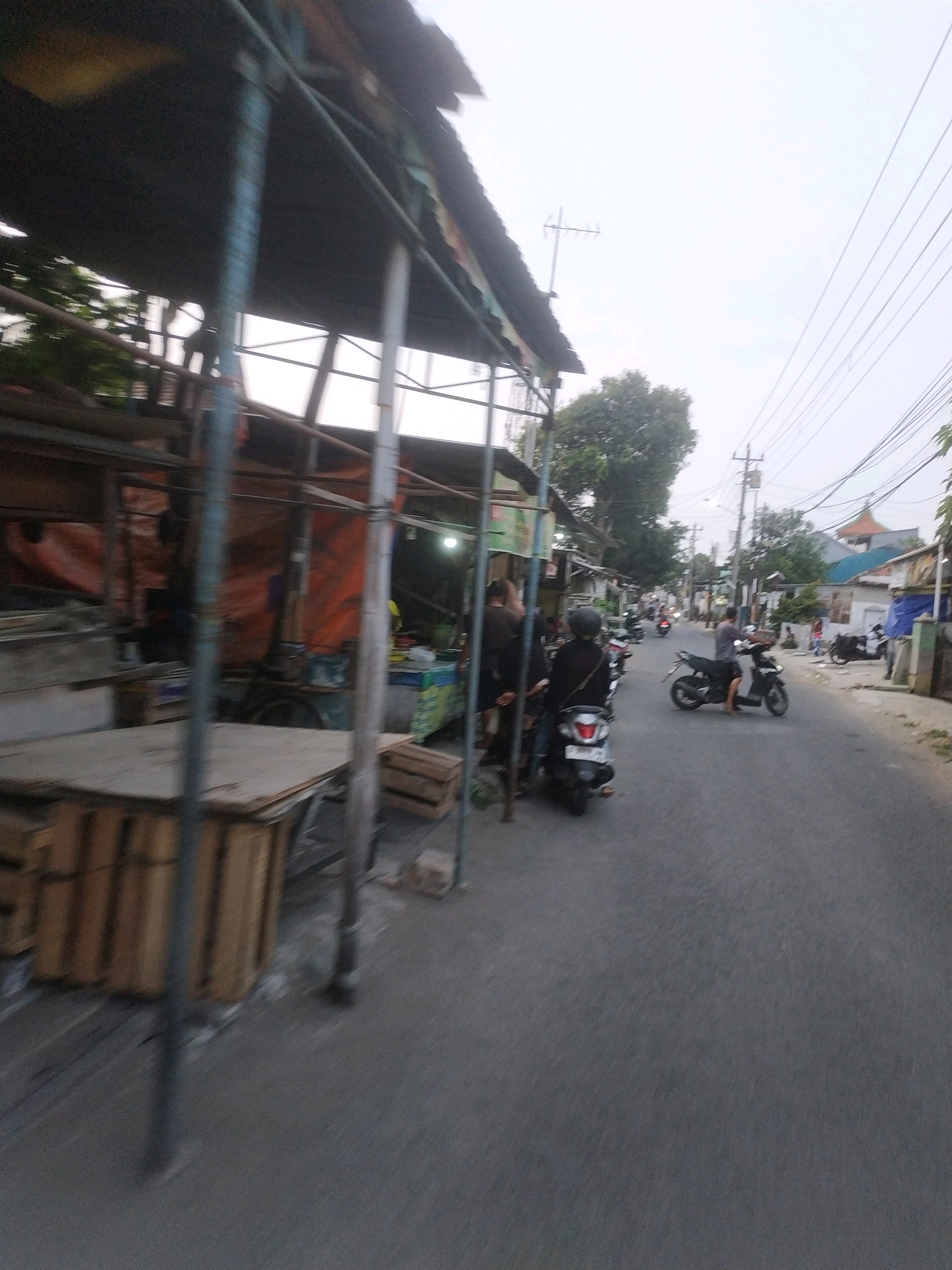 The image shows a street scene in Tegal, Indonesia, likely a local market or roadside stalls. In the foreground, there are several wooden structures, possibly part of temporary market stalls, covered with a weathered roof supported by metal poles. Some of these structures contain stacked wooden crates and what appear to be plywood sheets.
Further down the street, several people are visible. Two individuals are on a motorcycle, with one driving and the other seated behind. Another person is on a separate motorcycle, also in motion. On the left side of the street, near the stalls, two people are standing and observing.
The street itself is paved asphalt. In the background, there are various buildings with different architectural styles, including some with tiled roofs. Utility poles with electrical wires are prominent along the street. Vegetation, including trees, can be seen interspersed between the buildings. The sky is overcast, suggesting a cloudy day. The overall impression is a busy, everyday street life in a populated area of Tegal.