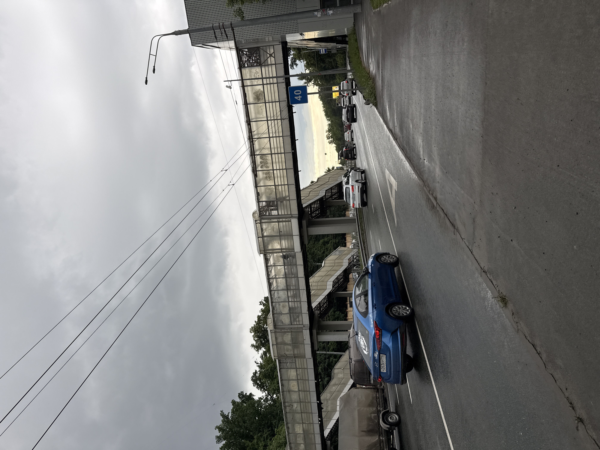 The image depicts a street scene in Kazan, Russia, with a focus on a pedestrian bridge overhead and traffic on the wet asphalt road below. The sky is overcast, suggesting a cloudy or rainy day. The pedestrian bridge is a prominent structure, made of glass and metal, with visible staircases leading up to it. The road is lined with trees and appears to be a busy thoroughfare, with several cars visible.
The activity taking place is vehicular traffic on the road, and people are likely using the pedestrian bridge, though none are clearly visible in the frame. Notable details include the wet road surface, indicating recent or ongoing precipitation. A speed limit sign showing "40" is clearly visible on the pedestrian bridge, indicating a speed restriction on the road. The time of day appears to be daytime, given the ambient light, but the cloudy conditions prevent a precise determination. The presence of a speed limit sign with Russian text and license plates on the cars suggests the location is in Russia.
There are no discernible emotions conveyed by the scene, as it is a candid shot of an everyday street environment. The image captures a functional urban setting with infrastructure for both pedestrians and vehicles. The overall mood is muted due to the overcast weather and the wet conditions.