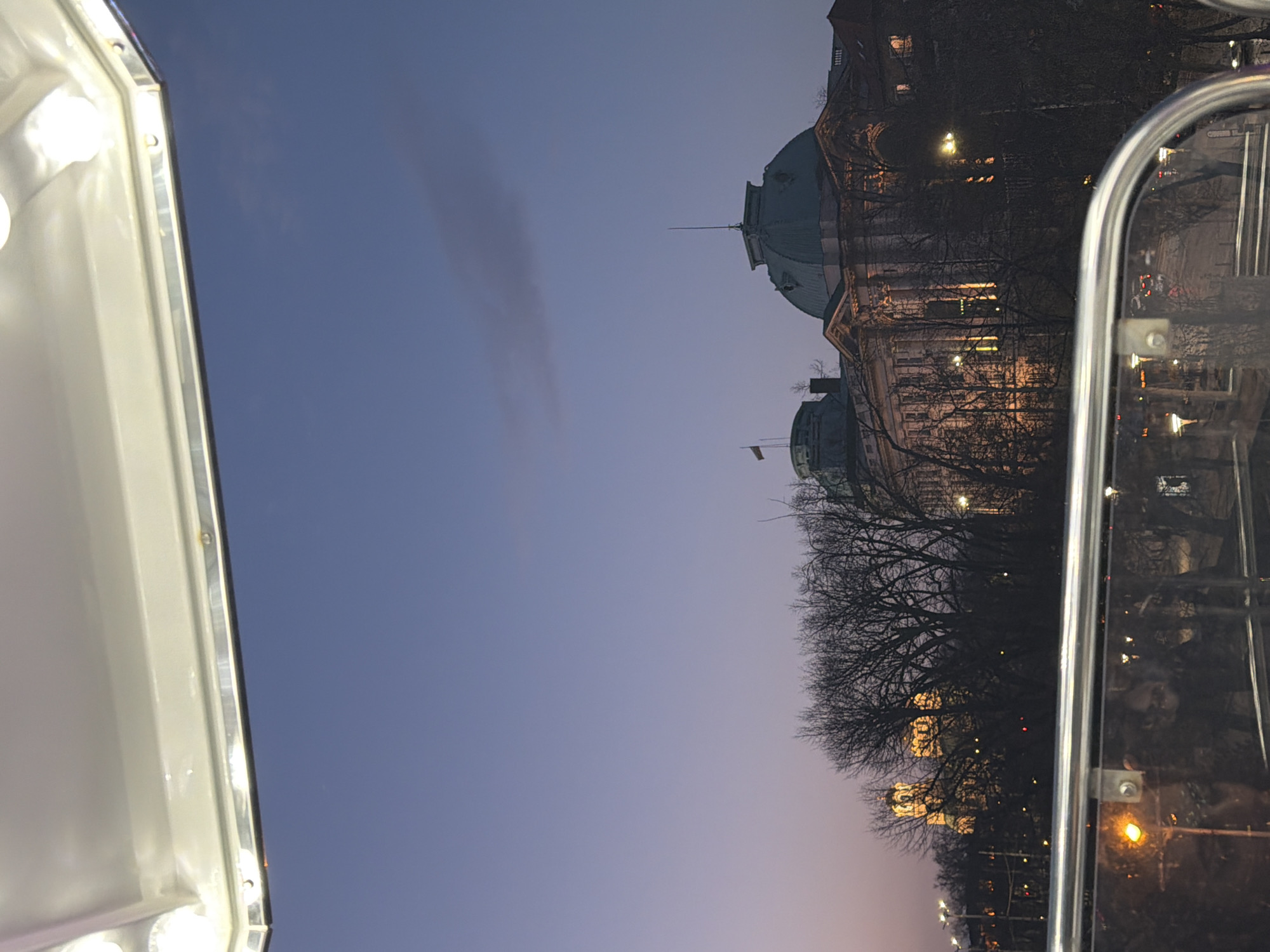 Captured from the inside of a vehicle, the image showcases Sofia, Bulgaria's cityscape against a dusk sky. The left foreground is dominated by a white frame with illuminated bulbs, contrasting against the dark sky. To the right, a blurred reflection in glass reveals glimpses of city lights and a possible face. The buildings stand silhouetted against the sky, interspersed with bare trees. The architecture features at least two prominent domes and lit windows.