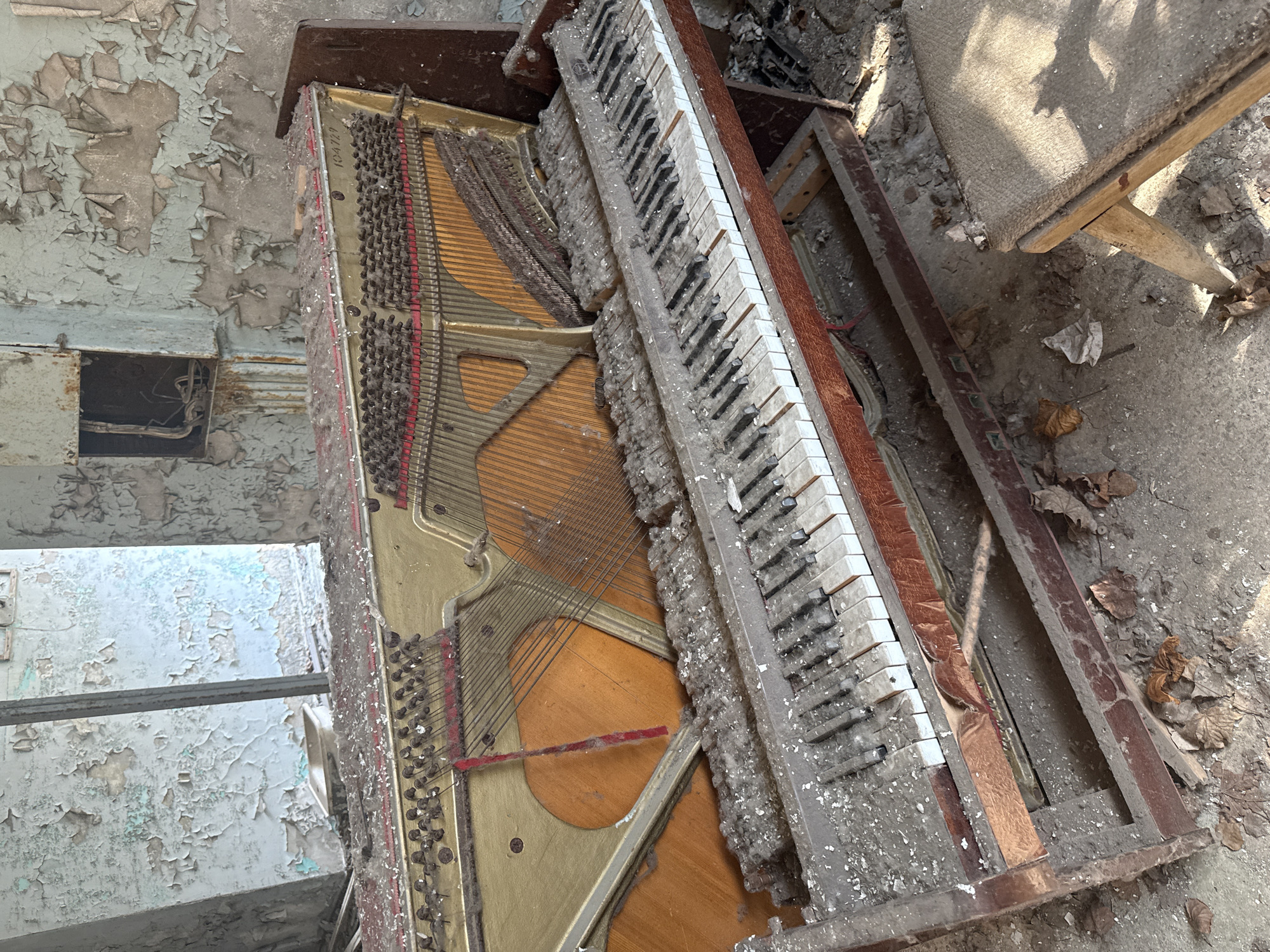 The image depicts a severely deteriorated upright piano in an advanced state of decay within an abandoned building, likely in Prypiat, Ukraine, given the prevalence of such scenes there. The piano's wooden frame is broken and covered in a thick layer of dust and debris. The keyboard is heavily soiled and some keys are missing or damaged. The internal components, including strings and hammers, are exposed and in disrepair. The surrounding environment shows signs of extreme neglect, with peeling paint on walls and debris scattered on the floor. A partial view of a broken chair is also visible, further indicating abandonment and disuse. There is no evidence of human presence.