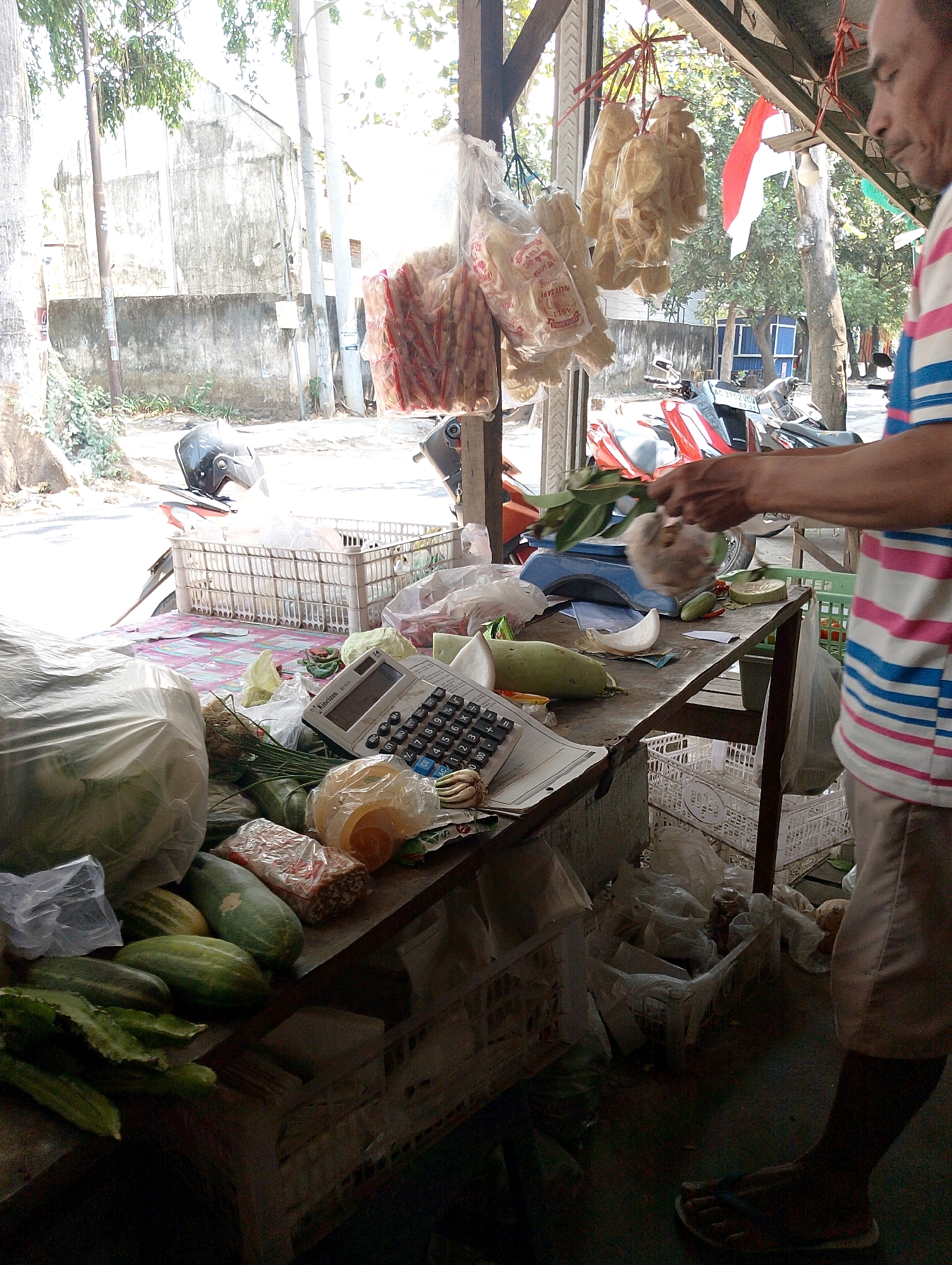 An outdoor market stall in Berbek, Indonesia, displays a variety of goods. In the foreground, a table is laden with fresh produce, including green beans, cucumbers, and what appear to be gourds. A calculator is visible on the table, along with various plastic-wrapped items. Hanging from the stall's roof are bags of what seem to be crackers or snacks. A person, partially visible from the side and wearing a striped shirt and shorts, is interacting with the produce. In the background, motorcycles are parked, and a street scene unfolds with buildings and trees. The overall impression is one of a bustling local market setting.