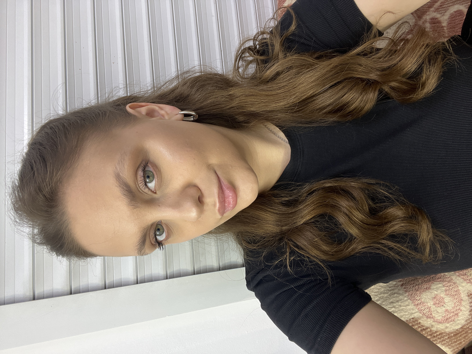 A young adult female is depicted in a frontal, chest-up close-up, looking directly at the camera with a subtle smile. Her medium-length, wavy brown hair is parted on the left and flows over her shoulders. She exhibits fair skin, light green eyes, and pink-toned lips. She is wearing a black, short-sleeved ribbed top, a thin silver-colored chain necklace, and rectangular silver-colored hoop earrings. Portions of a patterned textile, featuring cream, pink, and brown hues, are visible on the lower left and right, suggesting she is seated or leaning against it. The background consists of a stark white vertical wall section on the left, next to a white, horizontally slatted surface, consistent with a closed roll-up door or security shutter. The scene is located in Chaikovsky, Russia.
