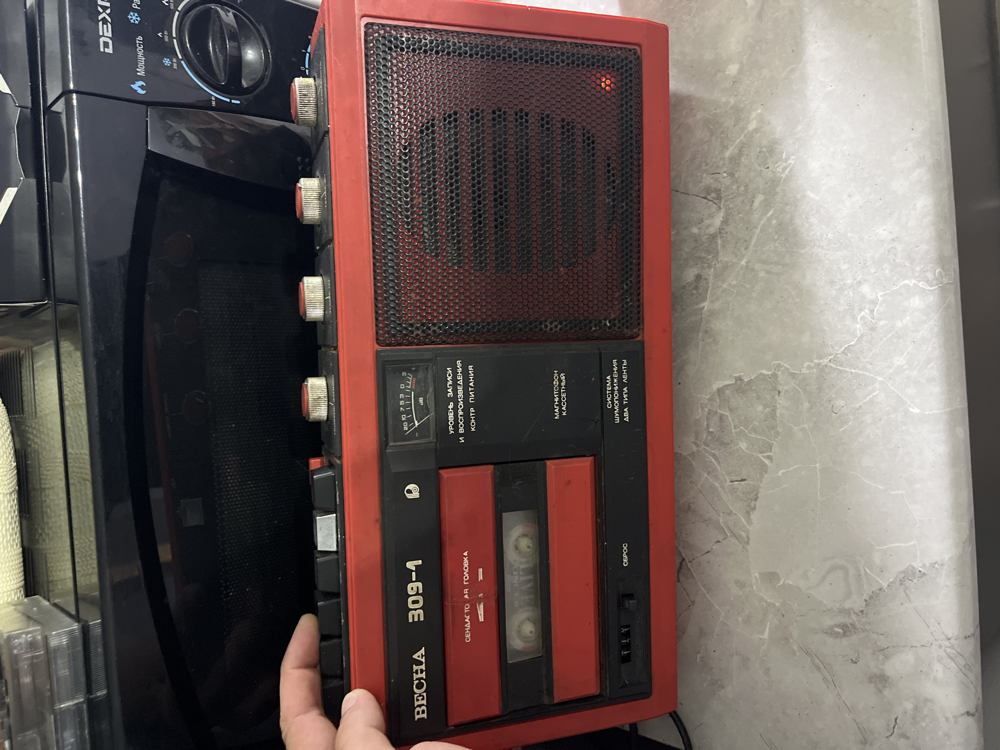 An overhead, medium shot captures a red portable cassette player, labeled "VESCHA 309-1," resting on a gray marble countertop. The cassette player has a prominent speaker grill on the right side, with a small red indicator light visible. To the left of the speaker, there is a cassette tape inserted in a red slot. Above the cassette slot, various control buttons and knobs are visible, including a slider and several round dials. A hand is partially visible on the far left of the image, near the buttons. In the background, a black microwave oven with the brand name "DEX" is visible, its door slightly ajar. The overall scene appears to be indoors, likely in a kitchen or workshop.
