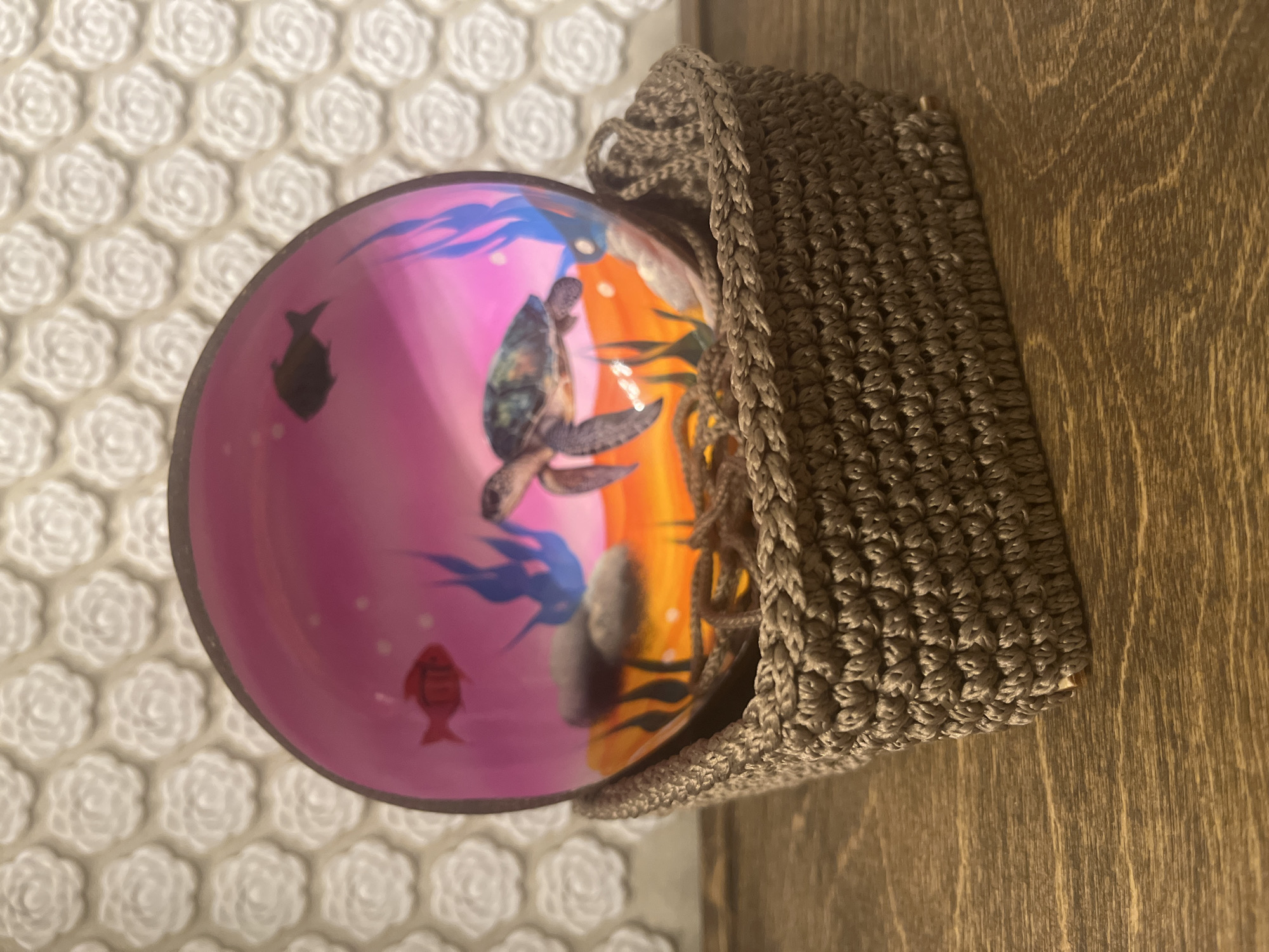 A high-angle close-up shot shows a bowl resting partially within a crocheted holder on a wood surface. The bowl has a pink background on the left with images of a sea turtle and fish. The right side of the bowl has an orange and yellow background with what appears to be coral. The crocheted holder is a neutral light brown color. The wood surface is a medium tone with visible grain. The background on the left is a white textured surface with a repeating floral pattern. There is no information about Novosibirsk, Russia, in the image.