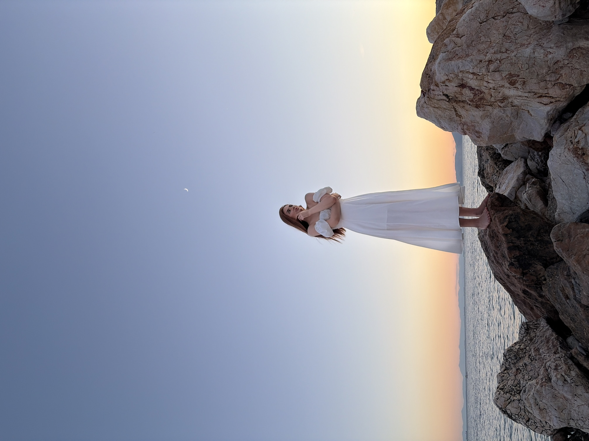 A young woman stands on large rocks near the sea in Paleo Faliro, Greece, at dusk. She is wearing a white, off-the-shoulder dress. The sky is a gradient of blue, transitioning to orange and yellow near the horizon. A crescent moon is visible in the upper portion of the sky. The sea is calm, with a distant coastline visible. The rocks are varied in size and texture, with some showing signs of erosion.