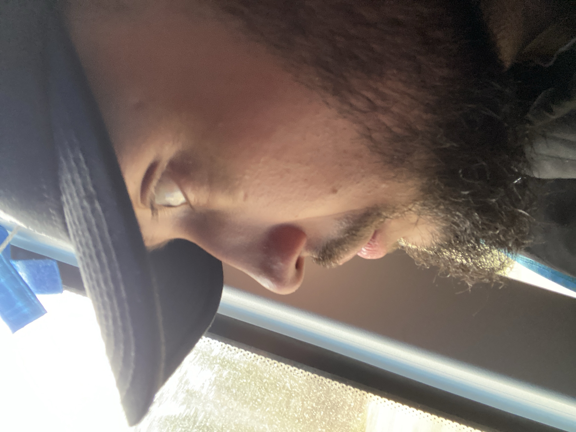 An upside-down close-up portrait shows the right profile of a person, likely male, from the forehead to the chin. The person has light skin, dark facial hair forming a beard and mustache, and dark hair. A portion of one eye, with a visible white sclera and iris, looks forward or downward. The individual is wearing a dark baseball cap, with its brim covering part of the forehead on the left side of the frame. A dark fabric, possibly part of a jacket or hoodie, is visible on the opposite side.
The setting appears to be inside a vehicle, possibly a car or bus, with the person looking out a window. The lower portion of the image (when oriented correctly) reveals a window frame and the bright, sunny view outside, showing what resembles green foliage or trees, slightly blurred. Above the window frame, a light-colored interior surface is visible. Some blue tape or material is affixed to the far left side, near the cap, suggesting a temporary fixture or repair.
The bright natural light streaming through the window indicates it is daytime, likely sunny. The person's expression appears neutral, suggesting observation or rest. There is no discernible text visible in the image. No specific visual cues confirm the provided location context of Bereznyaki, Russia.