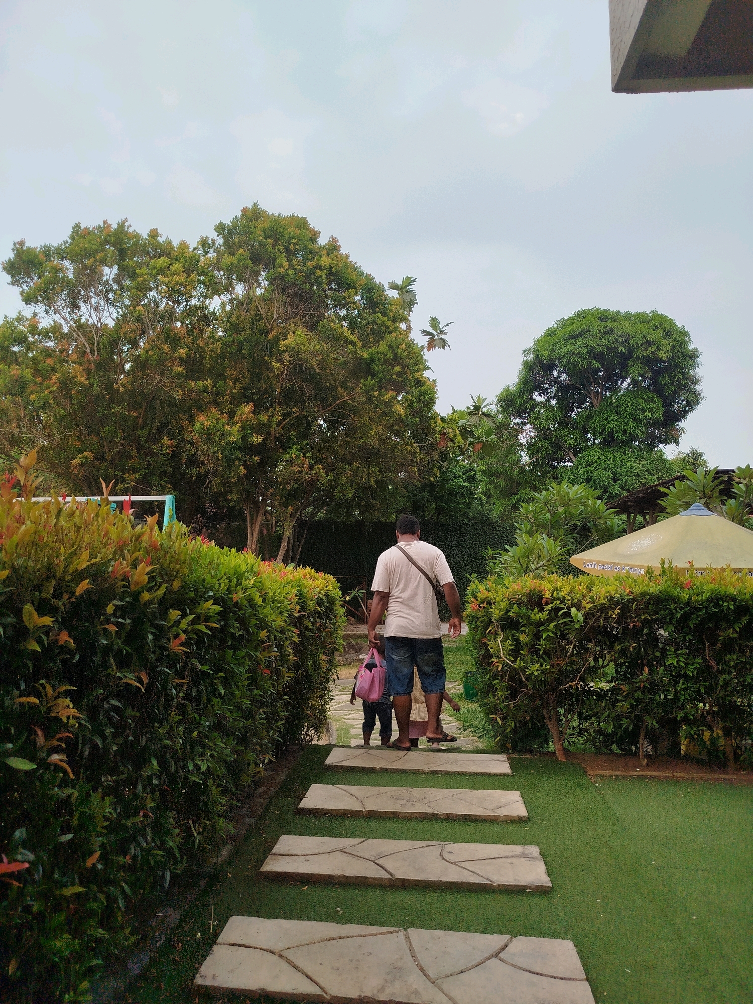 A man and a child are walking on a stone pathway in a park-like setting. The man is wearing a light-colored t-shirt and denim shorts, with a dark bag slung over his shoulder. The child, who is walking in front of the man, is wearing a pink backpack. They are surrounded by lush green hedges and trees. In the background, there is a large umbrella and more foliage. The sky is overcast. The scene appears to be a public park or garden, possibly located in Bugangin, Indonesia, given the surrounding vegetation and atmosphere.