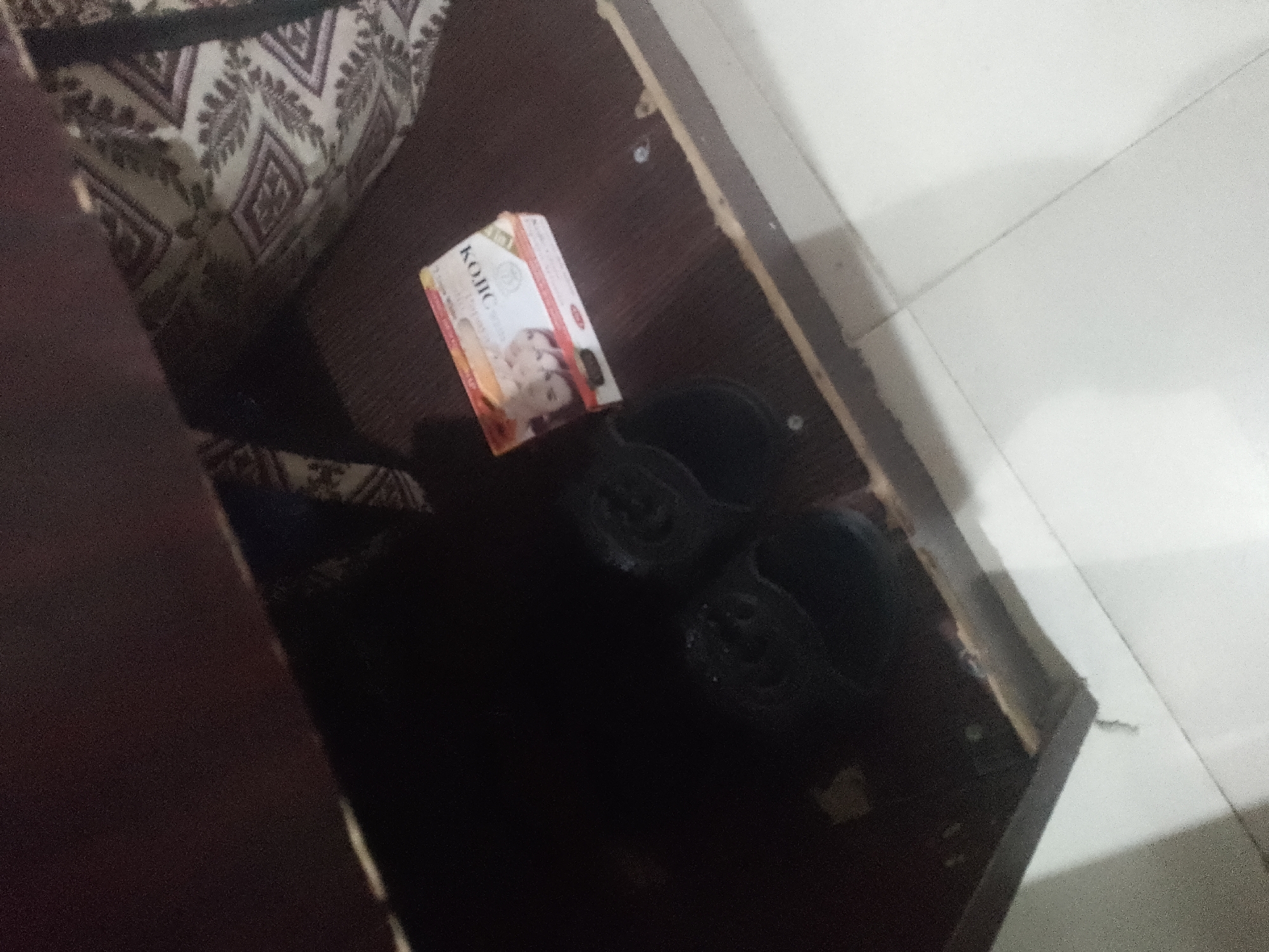 Inside a storage compartment, a box of Kojie San skin lightening soap sits on a dark brown surface. Two black slippers are placed next to the soap box. The compartment appears to be part of a piece of furniture, possibly a bed frame or sofa with a patterned fabric visible in the upper left corner. The background is a light-colored tiled floor.