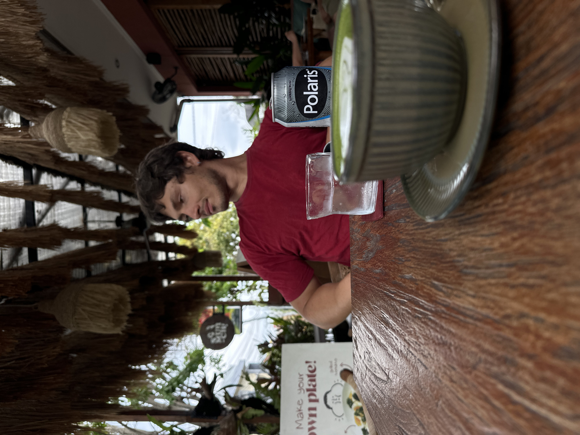 A young adult male with curly brown hair is seated at a wooden table in an outdoor seating area. He is wearing a red t-shirt and looking to his right. In front of him on the table is a glass of water, a silver can of "Polaris" sparkling water, and a bowl of green liquid, likely matcha. Behind him, a sign reads "Make your own plate!" The background shows foliage and a thatched roof structure. The scene appears to be a cafe or restaurant in North Kuta, Indonesia.