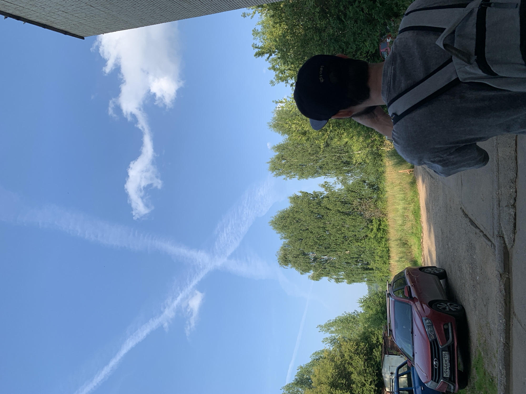 A person wearing a grey t-shirt and a black baseball cap is standing in a courtyard and looking at the sky. They are wearing a grey backpack. In the background, there are trees, a red car parked on a gravel surface, and a building. The sky is blue with contrails forming an "X" shape and some clouds. The location is identified as Chaikovsky, Russia, likely a residential area given the presence of the car and building.