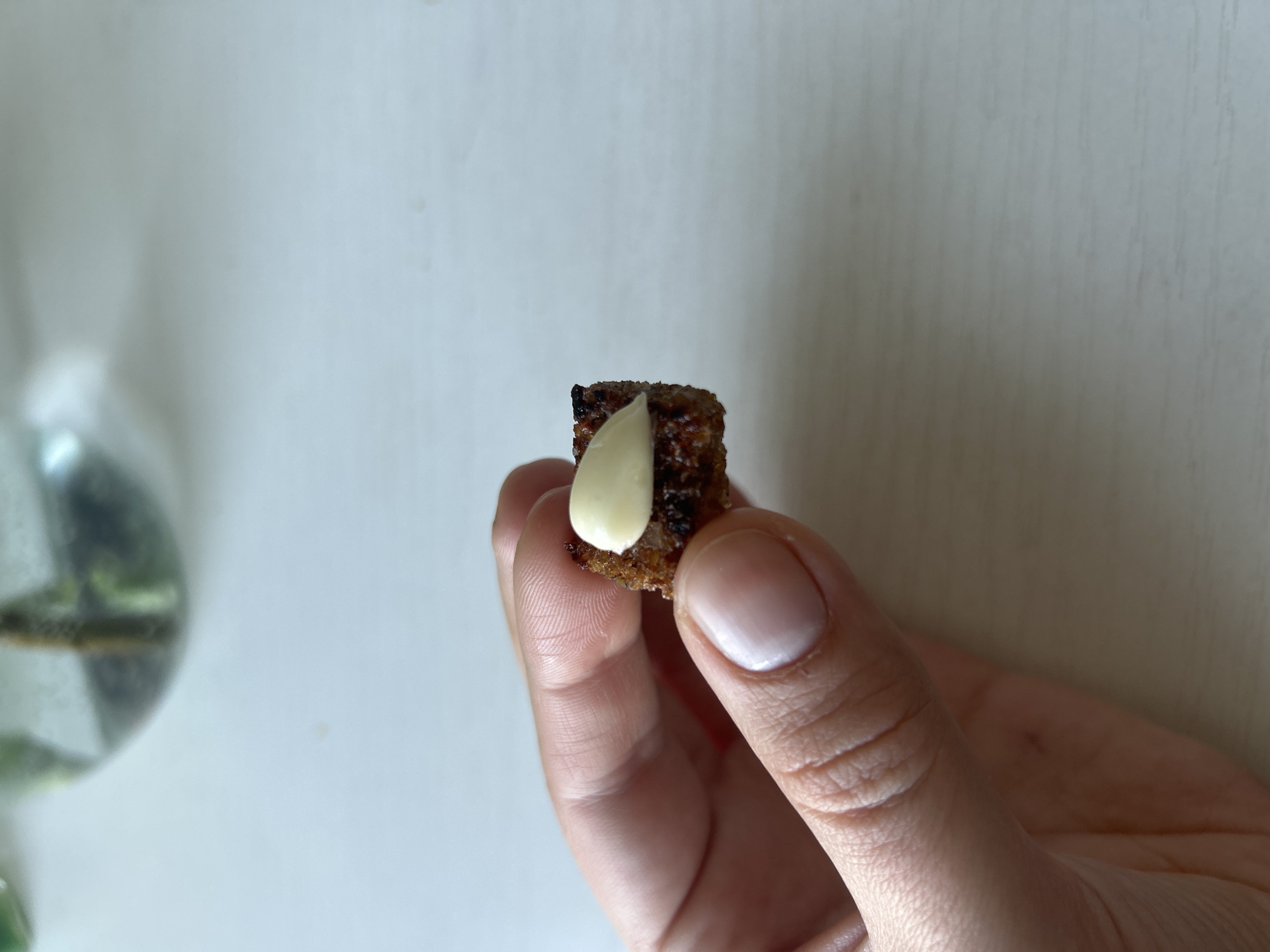 A person holds a small, dark brown crouton topped with a dollop of creamy white substance, possibly cheese or a similar spread. The person's hand has light skin, manicured nails, and visible lines. The background is a white surface with a blurred clear glass object containing green stems and water to the left. The location is unknown.
