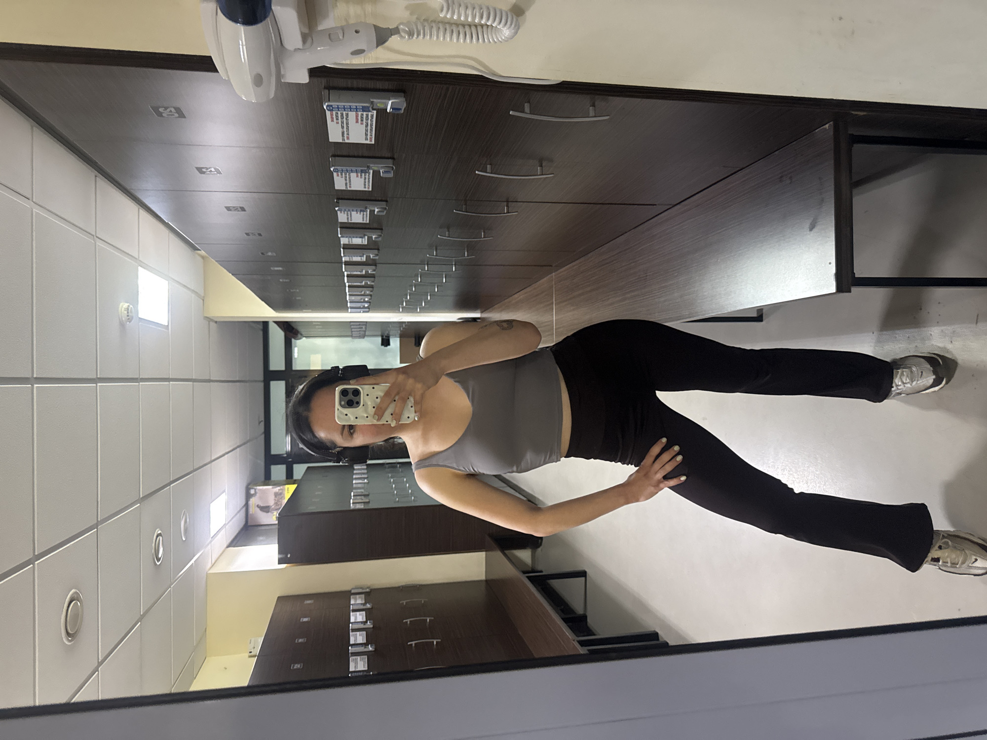 A person is taking a mirror selfie in a room with lockers. The person is wearing headphones, a grey top, and black leggings. They are holding a phone with a white case and taking the photo. The lockers are dark brown with silver handles. There is a white wall on the left side of the frame with light fixtures. The top of a medical device is visible above the lockers. The location is unknown, but it is unlikely to be in Burgas, Bulgaria, as there is no visible indication of this.