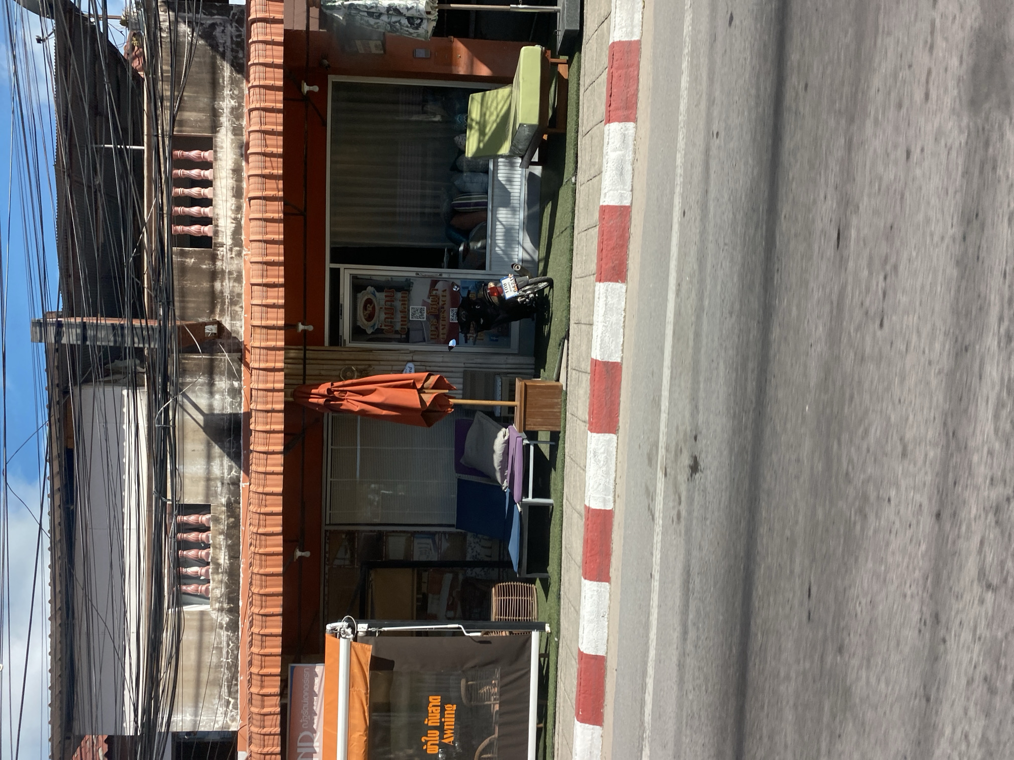This is a horizontal, eye-level, medium shot of a storefront in Baan Hua Thanon, Thailand. The storefront has an orange awning and displays furniture outside. The building appears to be two stories, with a balcony on the second floor. There are many power lines strung across the top of the frame. The foreground is dominated by a street with a red and white striped curb. A motorcycle is parked just inside the doorway of the shop. There are two lounge chairs outside the shop, one dark blue and one purple, with a pillow on the purple one. To the right is a green lounge chair with a cushion. An orange umbrella is folded and leaning against a wooden stand. The shop window is covered with blinds and shows a display of pillows. The awning on the left side of the frame has text in Thai and English that reads "Awning". The overall scene suggests a commercial area in a warm climate.