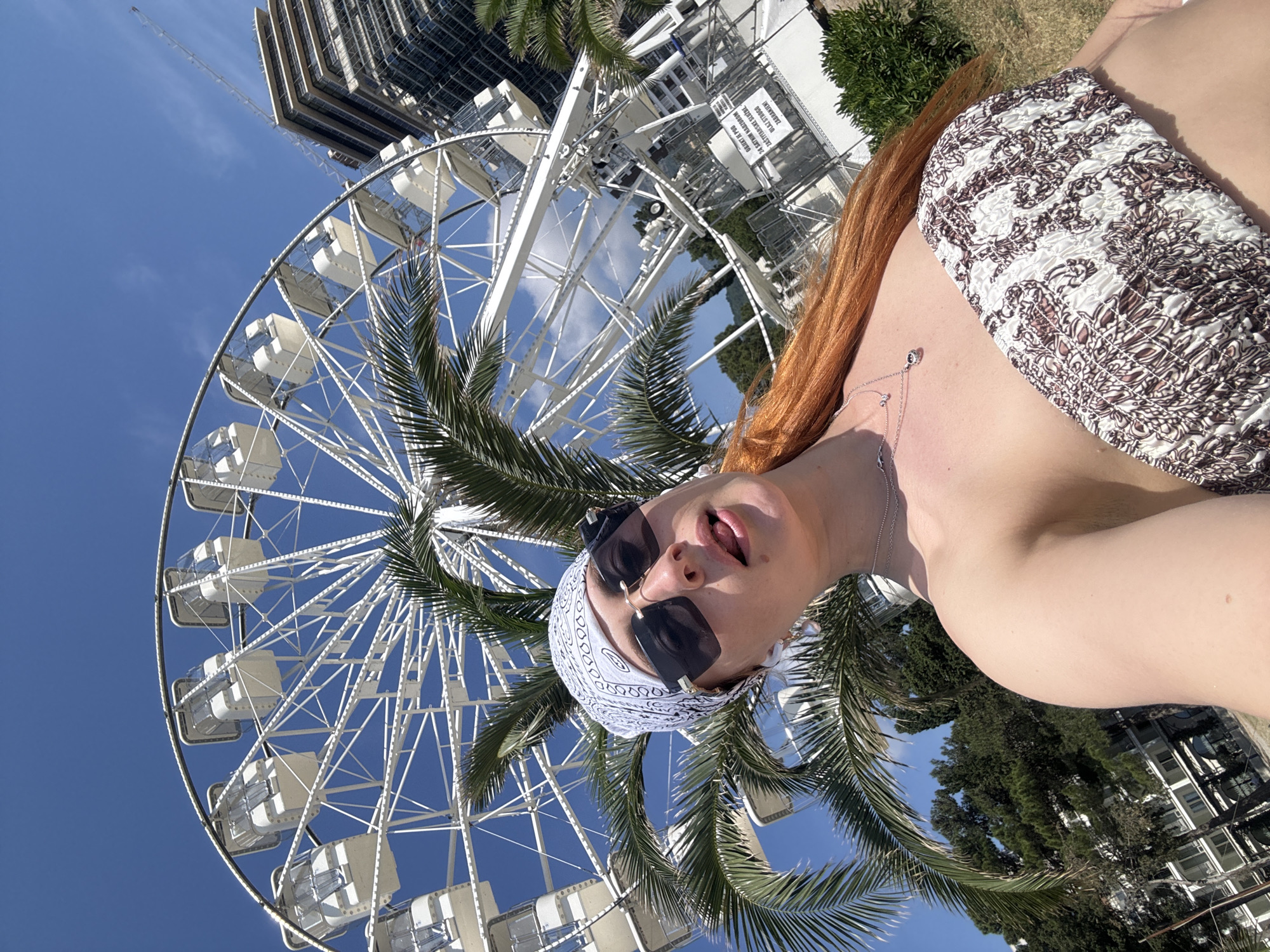 The image is an angled selfie taken in Budva, Montenegro, against the backdrop of a large Ferris wheel, palm trees, and a building under construction.
The subject is a fair-skinned person with long red hair, wearing a white and patterned strapless top, a white bandana, and dark sunglasses. They are positioned on the right side of the frame, facing the camera with their mouth slightly open. A delicate necklace with a pendant is visible around their neck.
Behind the subject is the Budva Ferris wheel, partially obscured by palm tree foliage. The sky is clear and blue. The building under construction is located at the top left of the image.
The setting appears to be an outdoor area near the Ferris wheel, possibly a park or tourist attraction. The image suggests a sunny day.