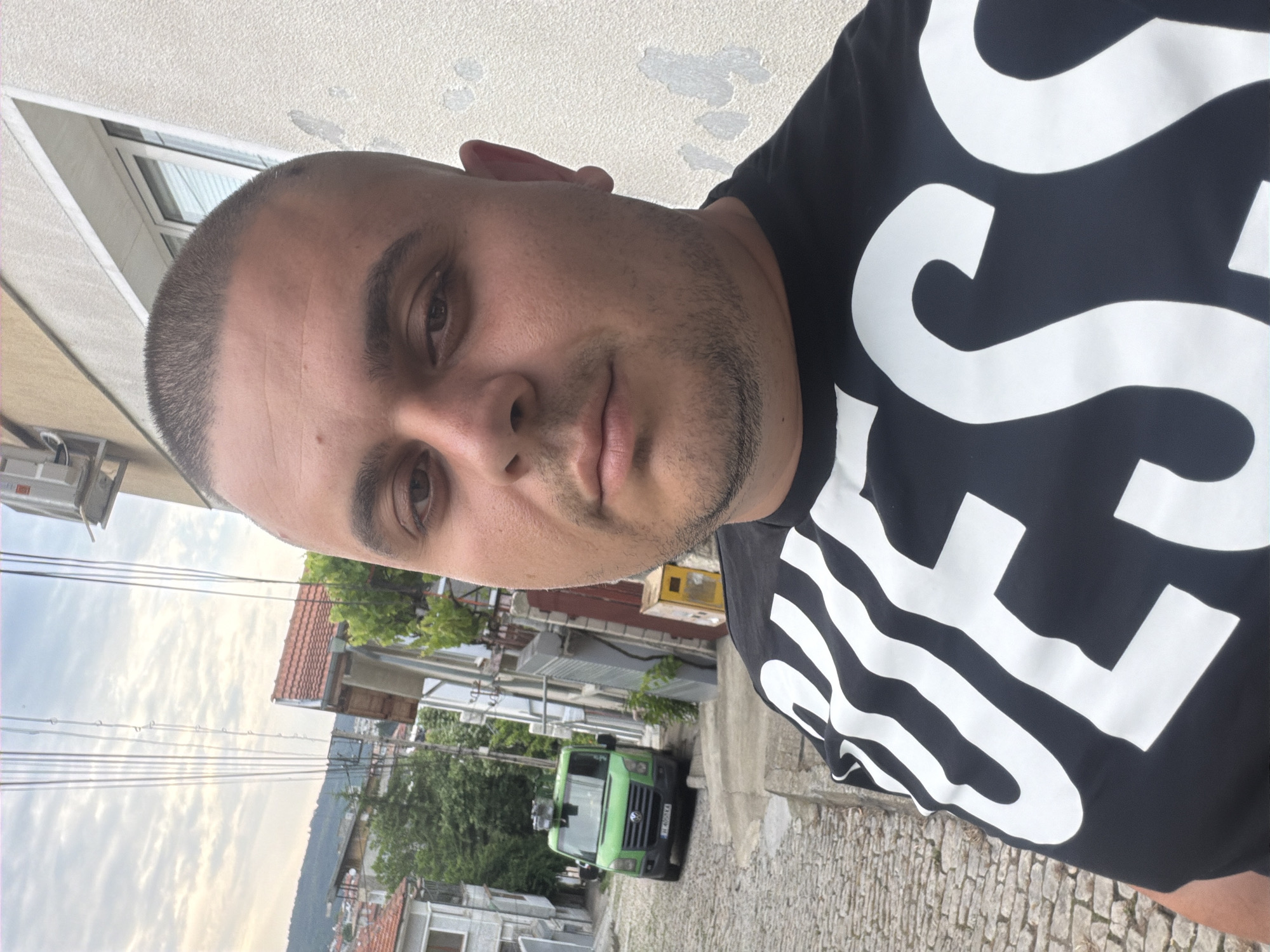 A detailed, direct-angle selfie captures an adult male with a buzz-cut hairstyle and stubble, wearing a black t-shirt emblazoned with "GUESS" in large white letters. His expression is neutral as he looks directly into the camera. The setting is an outdoor, urban street environment, characteristic of many Bulgarian towns, including areas within Shumen.
The street features cobblestone pavement and slopes downwards from the immediate foreground. On the left side of the street, portions of residential buildings with light-colored facades are visible. A green Volkswagen Crafter van is parked on the cobblestone street; its front license plate, "B 4051 KA", is clearly legible, indicating registration in Sofia, Bulgaria.
To the right of the subject, a light-colored building with peeling paint and a visible window forms the immediate background. An external air conditioning unit is mounted high on this building's wall. Below it, a yellow utility box is affixed to the wall. The background beyond the street reveals a densely built residential area with various buildings featuring tiled roofs, interspersed with trees and greenery. Multiple overhead utility wires traverse the scene against a partly cloudy sky. Distant hills or mountains are visible on the horizon, consistent with the geography of Shumen.