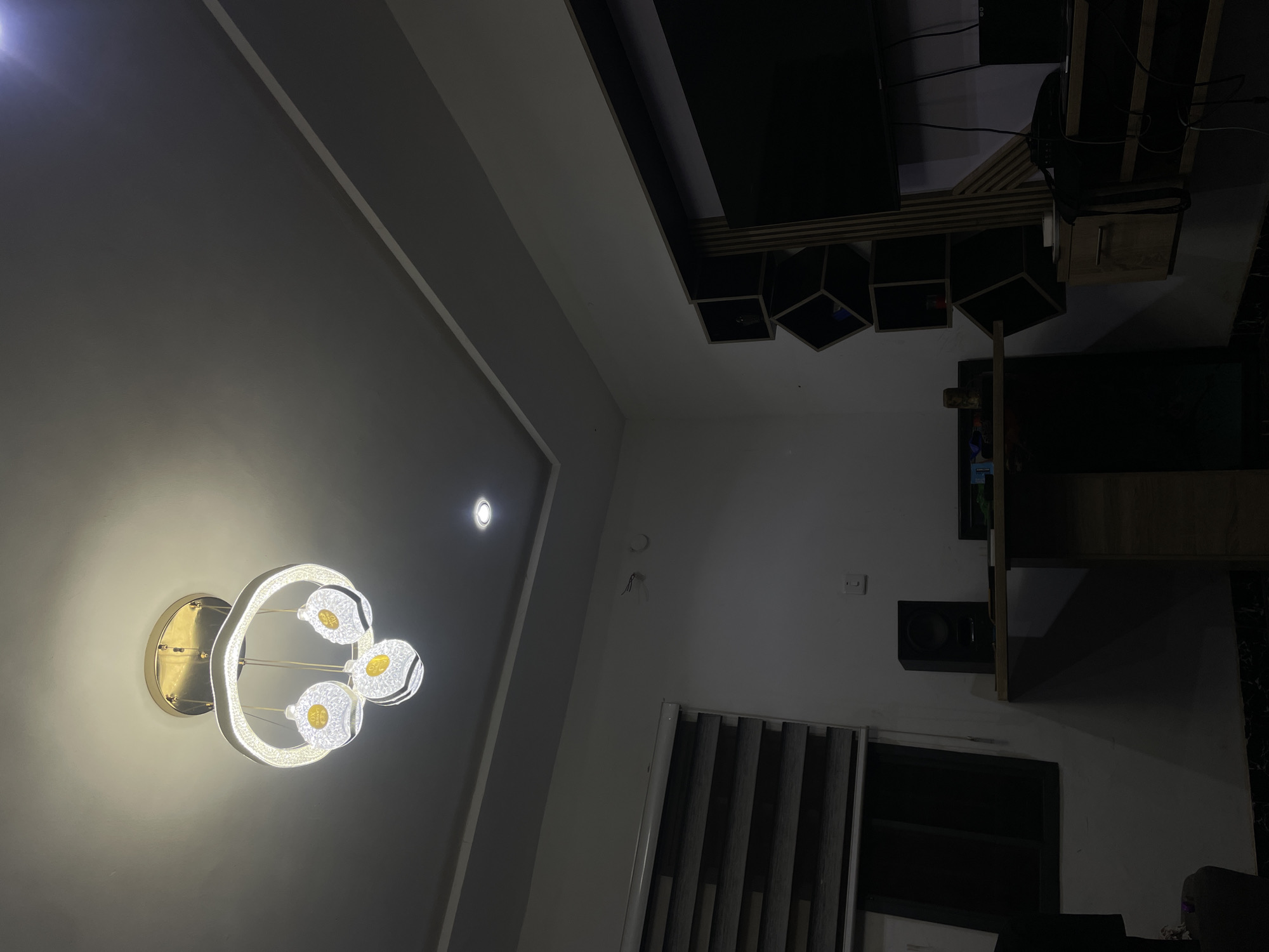 The image depicts the interior of a dimly lit room, likely a living room or entertainment area. Dominating the upper portion of the frame is a modern chandelier with a circular frame and three spherical light fixtures, casting a warm glow. The ceiling features recessed lighting.
On the left side of the room, a window is covered by a modern blind with horizontal slats, alternating between dark and light shades. Below the window, a black speaker sits on a wooden desk.
The right side of the room showcases a wall-mounted entertainment unit with a dark television screen. Adjacent to the TV are several black cube-shaped shelves, some of which are empty and others containing small, indistinct objects. The wall is painted a light color.
The overall ambiance is quiet and private. The scene appears to be a snapshot of a domestic space, likely within a residential building in Abuja, Nigeria, given the context provided. No people are visible in the image.
