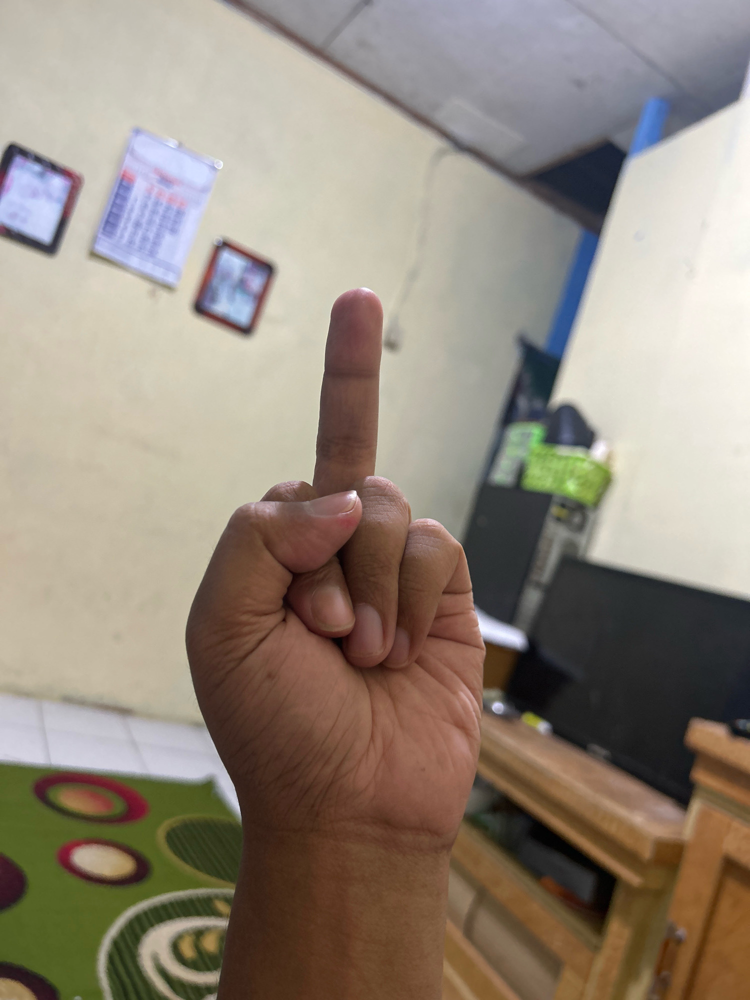 A close-up shot focuses on a person's right hand, specifically their middle finger extended upwards, with the index finger curled down and thumb against it. The background suggests an indoor setting, possibly a room, with a wall painted in a light yellow hue. Several framed pictures or calendars hang on the wall to the left of the hand. A wooden cabinet with drawers and doors is visible in the background to the right, topped with a dark rectangular object, likely a television. A green rug with an abstract pattern is present on the floor in the lower left portion of the image. The ceiling is visible in the upper part of the frame. The image was taken at an indoor location in Sukanagara, Indonesia.