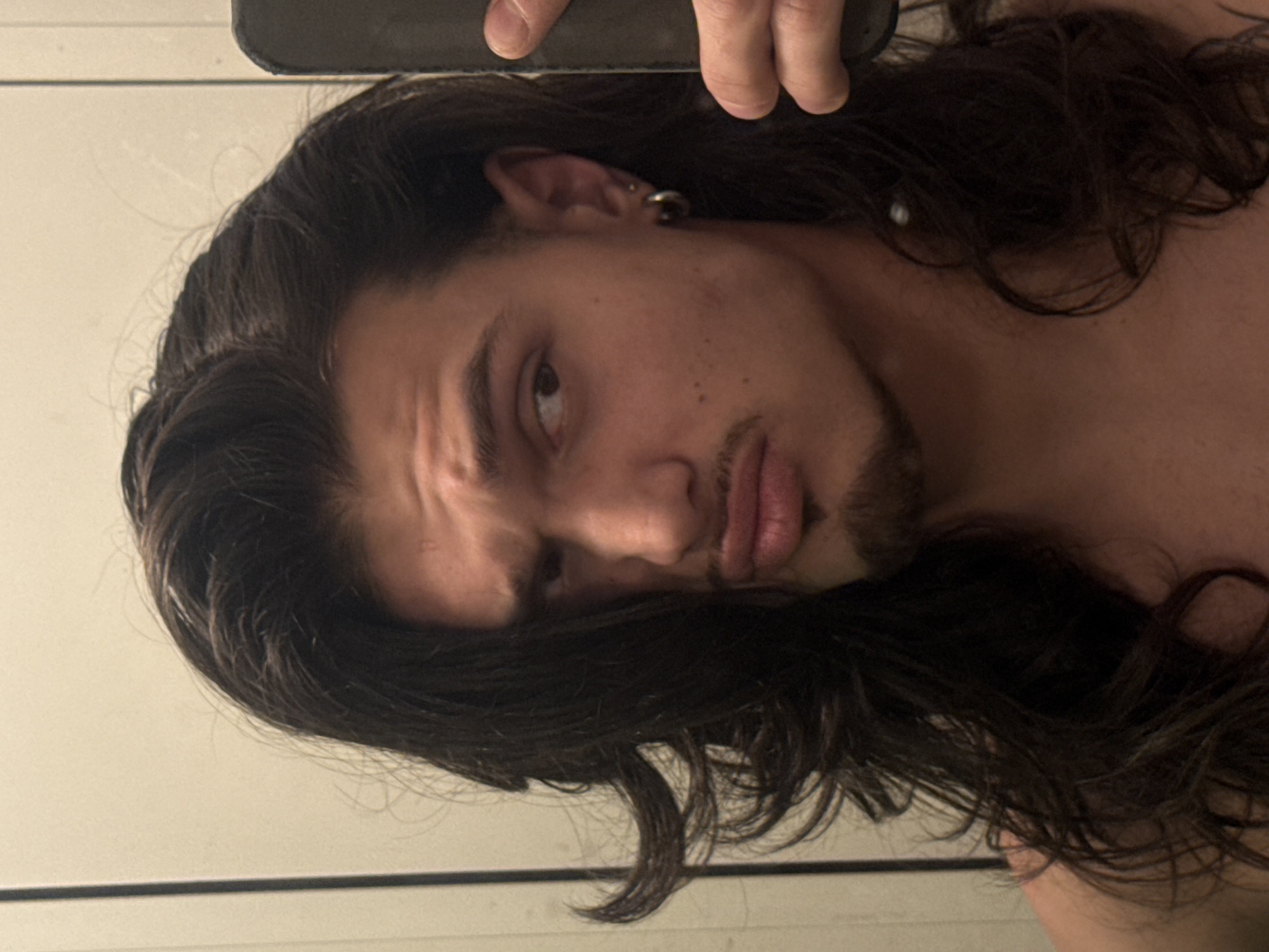 A young adult male with long, dark, wavy hair and a goatee is looking towards the camera with a pouty expression. He is wearing a hoop earring in his left ear and a smaller stud earring in his right ear. He is holding a dark-colored smartphone in his right hand, partially obscuring the right side of his face and body. The background appears to be a plain, light-colored wall, possibly in a bathroom due to a vertical reflection line that could indicate a mirror or door frame. The image does not contain any information that would suggest a location in Sofia, Bulgaria.