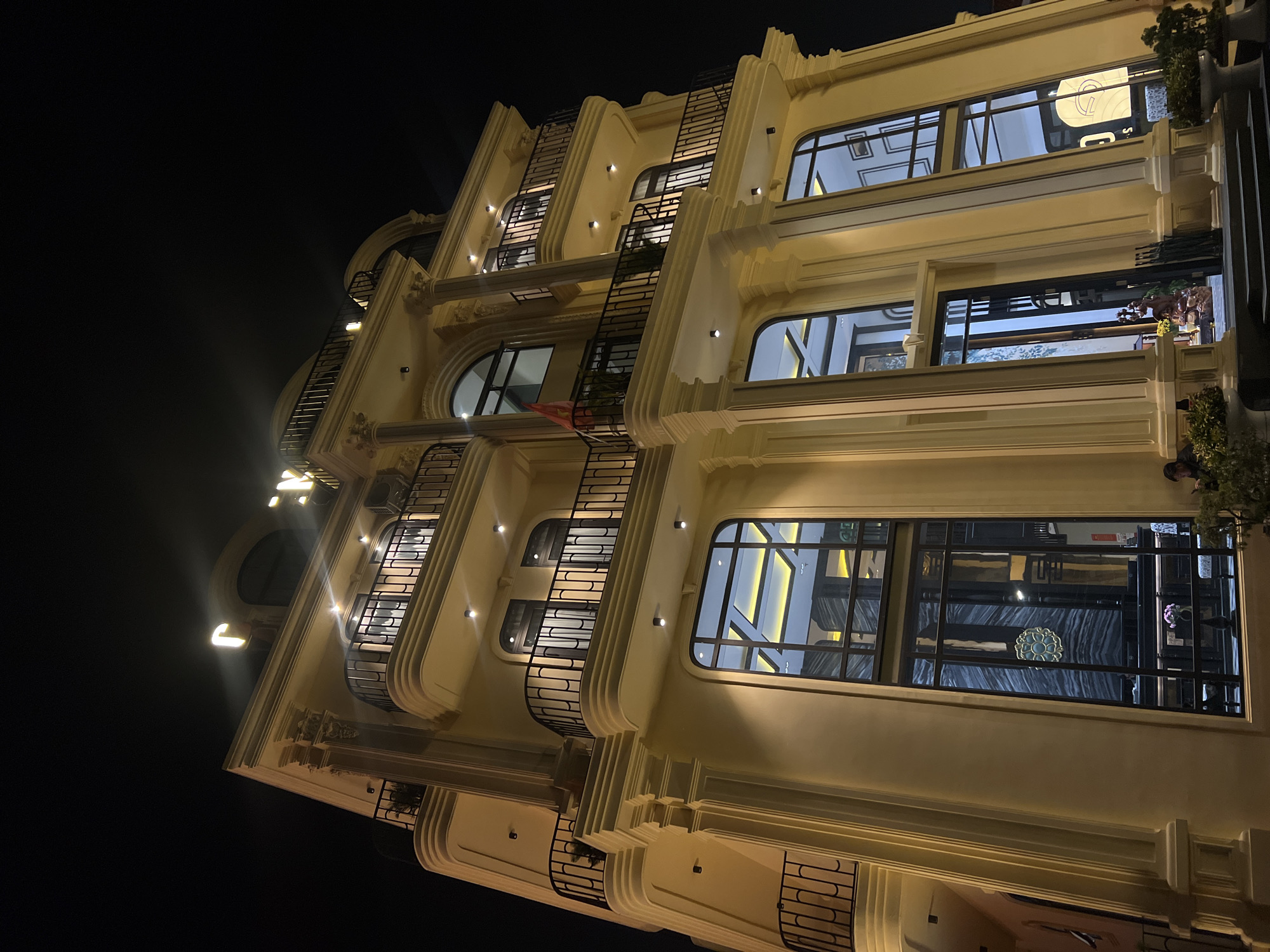 A low-angle photograph shows a multi-story building at night. The building has a pale yellow facade with arched windows and black wrought-iron balconies and fire escapes. The windows are lit from within, revealing interior architectural details. The building’s architectural style features stepped layers and ornate details along the cornices. The surrounding sky is dark, suggesting a nighttime setting. The location is Da Lat, Vietnam.