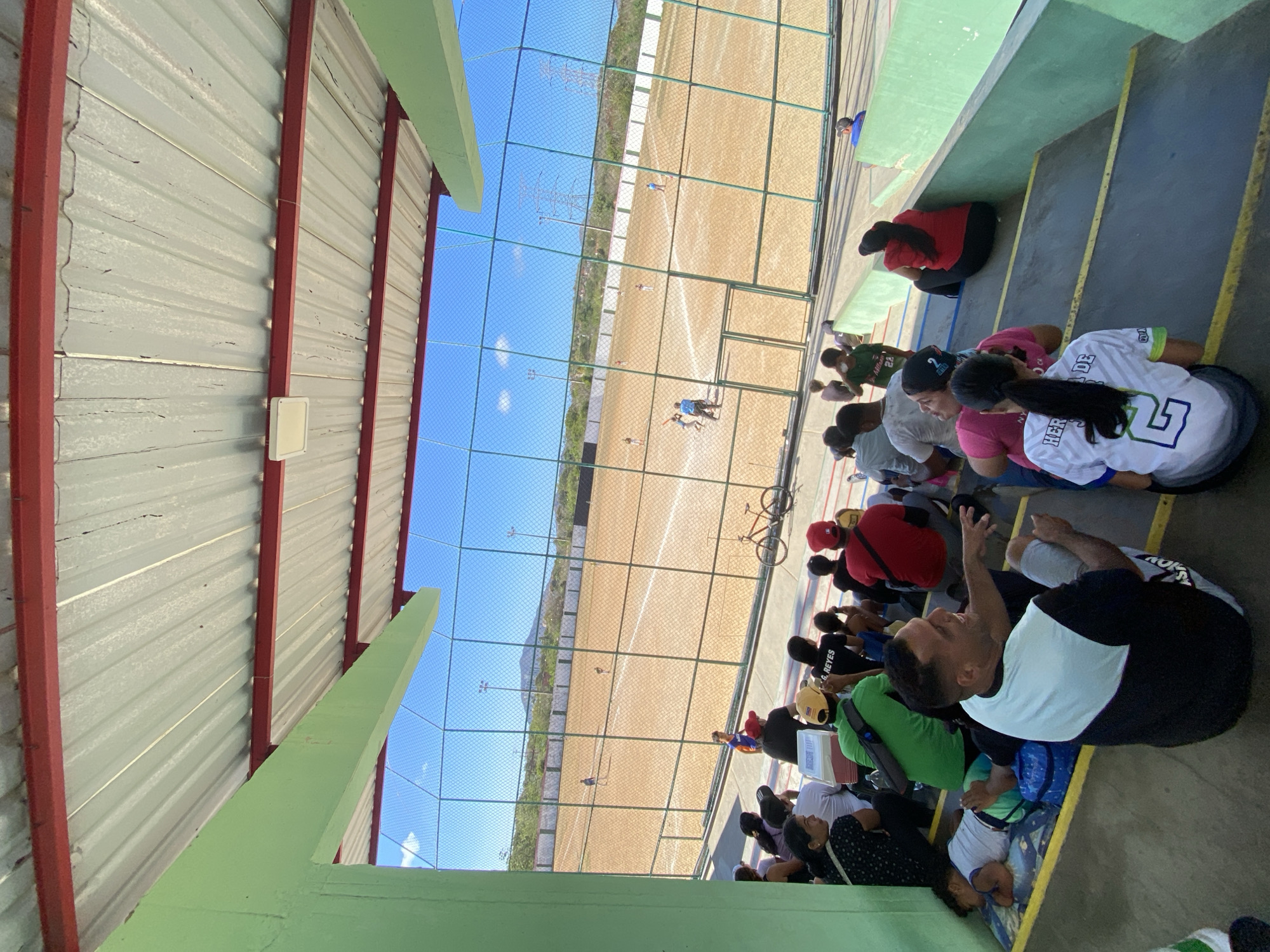 The image shows a group of people seated on stadium-style seating, likely watching an event on a running track visible through a chain-link fence. The track is tan and bordered by green lines. In the background, there are mountains and a clear blue sky. The viewing area appears to be under a metal roof supported by green-painted beams. The people are dressed in casual clothing, and some are wearing athletic jerseys. The location is the city of Juan Griego, Venezuela.