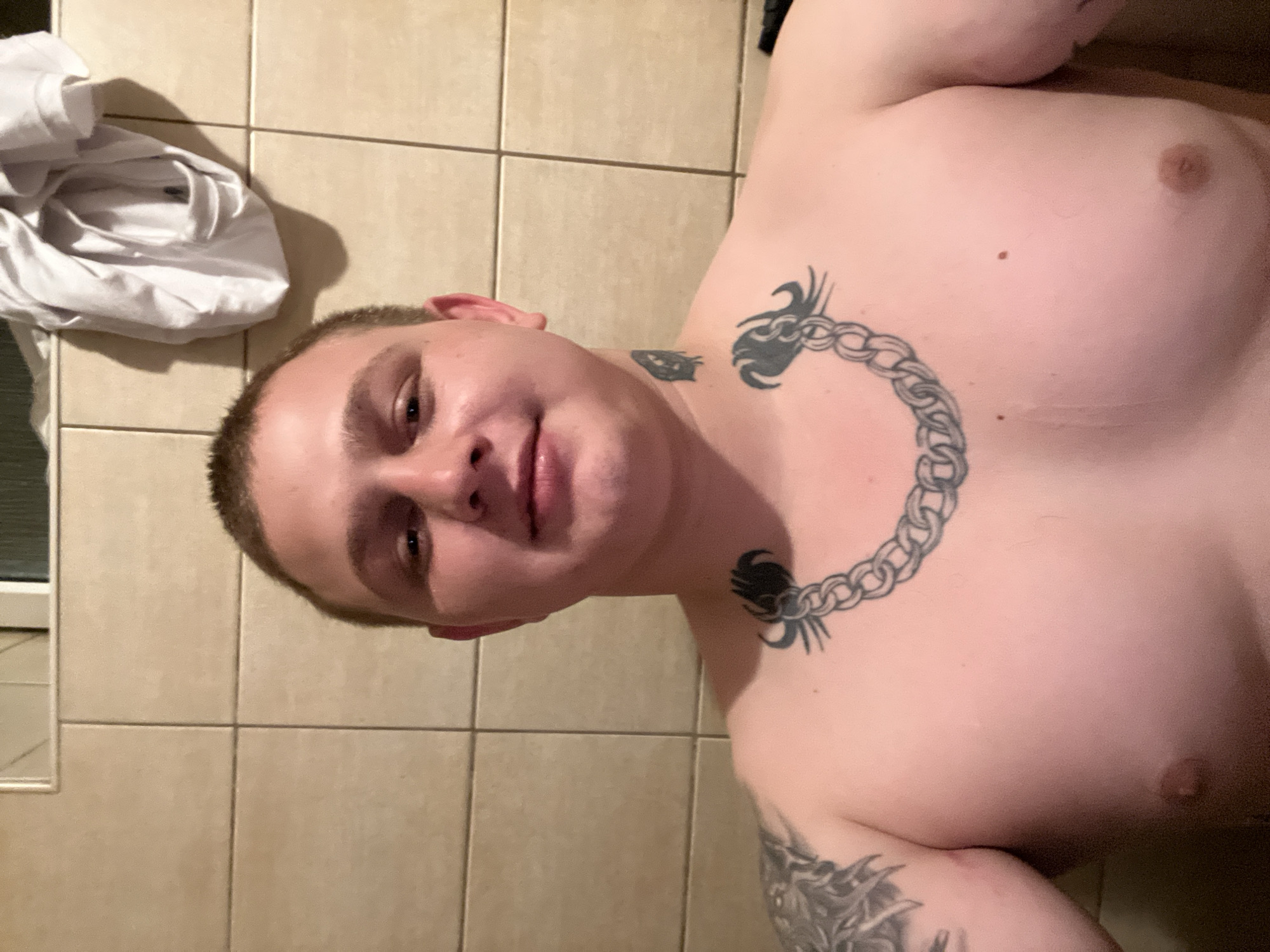 A shirtless man with short brown hair and a light complexion is pictured in a bathroom. He has several tattoos: a large one on his left arm and shoulder, a chain design around his neck, and a small one on his throat. The bathroom has beige tiled walls. A white shirt is draped over the tiles in the background. The image is a selfie taken from the chest up. The location in Pilsen, Czechia is not specified in the image.