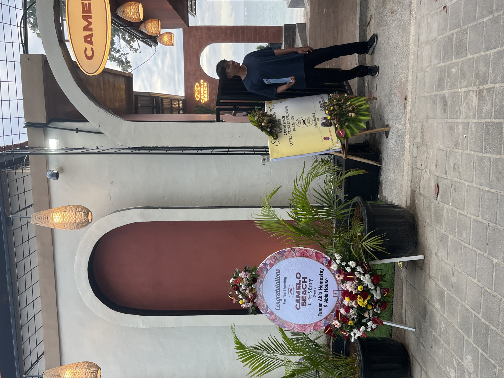 A young man stands in front of a building in Kuta, Indonesia, holding a blue folder. To his left, a circular floral arrangement congratulates the opening of "CAMELO BEACH Coffee & Eatery." Next to that, a yellow easel displays a sign announcing the grand opening of "CAMELO BEACH COFFEE, EATERY & CAMEL RIDING" on June 18, 2026. The building features arched doorways, a sign with the name "CAMEL," and hanging woven lamps. Plants in black pots are visible, and the ground is paved with gray cobblestones. In the background, the ocean can be seen through an archway. The sky is overcast.