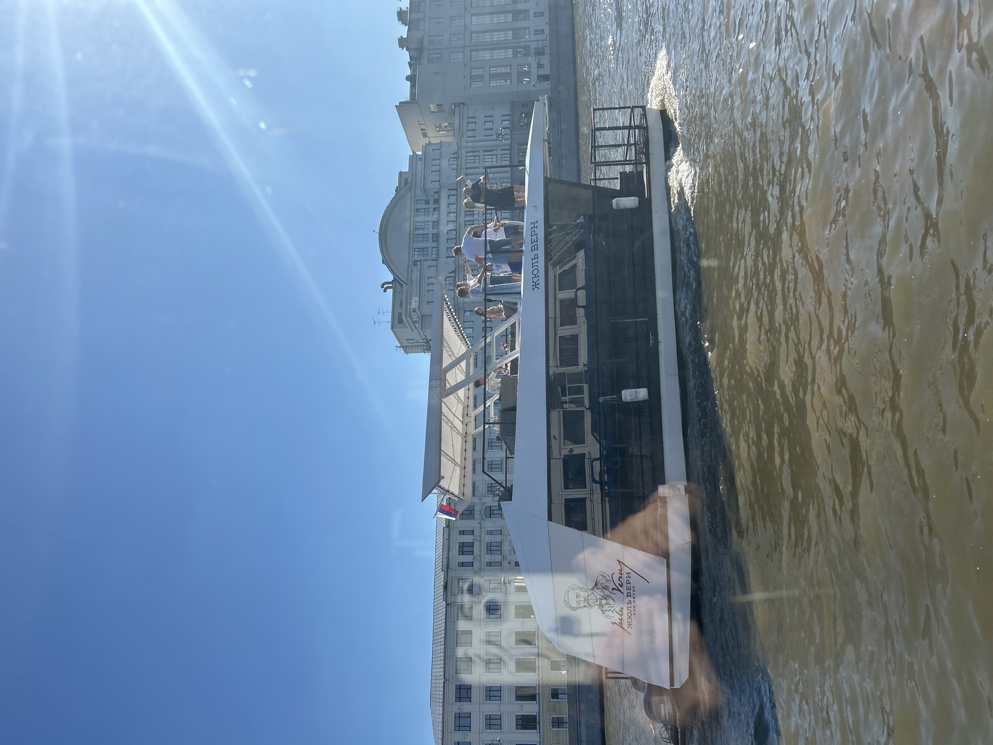A modern white and black boat named "Jules Verne" cruises on a body of water, likely a river or canal in Moscow, Russia. The boat features a portrait of Jules Verne and Russian text. Several people are visible on the upper deck of the boat, some appearing to be conversing or enjoying the ride. In the background, several large, multi-story buildings with light-colored facades line the waterfront, characteristic of Moscow's architectural style. The sky is clear and blue, with the sun casting a lens flare onto the image, suggesting a bright, sunny day. The water is somewhat murky, with ripples and waves indicating the boat's movement. The overall scene depicts a leisurely boat trip in an urban setting.
