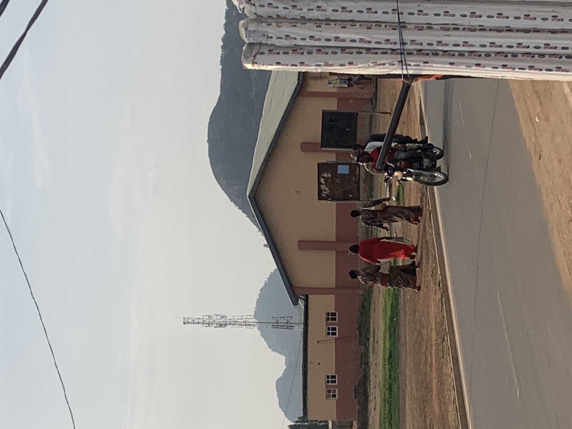 A daytime shot of a street in Sabojibi, Nigeria, featuring a group of people walking alongside a road next to a building. A motorcycle with two riders is parked on the road. The building has a tan and brown color scheme with several windows visible. In the background, a mountain and a communications tower are visible. Bundles of materials are stacked atop the building. An electrical wire runs along the left side of the image.