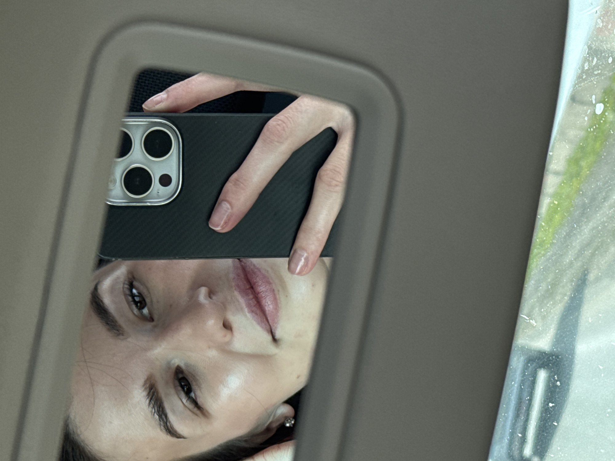 A woman is reflected in a car visor mirror, holding a smartphone with a multi-lens camera. The phone is black with a textured case. Part of the interior of the car, including the visor and a side window, are visible. The environment outside the window is blurred. The city of Noviy, Russia is not detectable in the image.