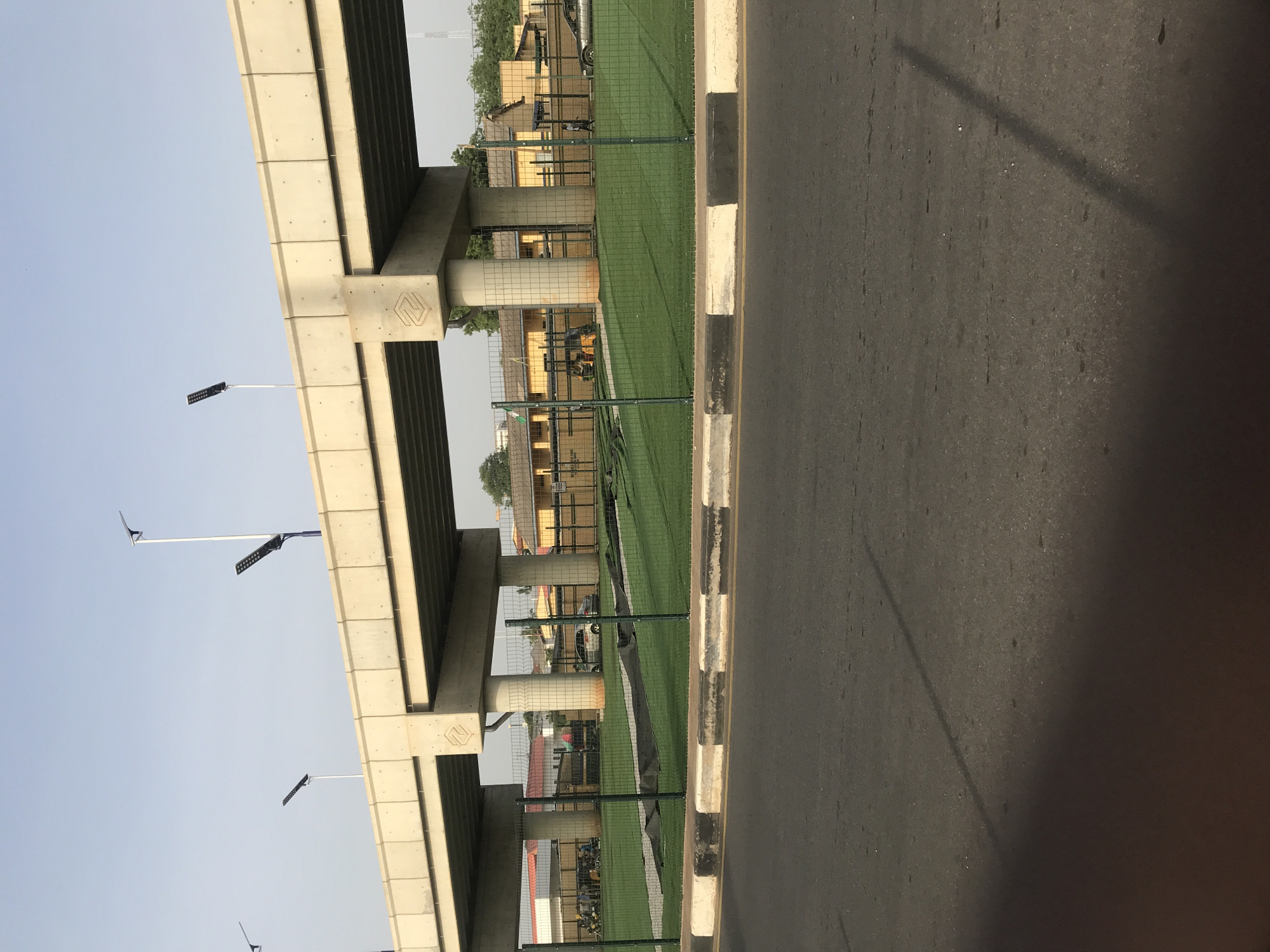 The image depicts a street scene in Mandate Estate, Ilorin, Nigeria. An elevated concrete overpass dominates the left side of the frame. Several light poles are positioned along the overpass. To the right of the overpass, there is a fenced area with artificial green turf. Behind the fence, there are buildings with yellow and brown walls. A concrete curb separates the fenced area from the paved asphalt road that occupies the right side of the image. The sky is light blue.