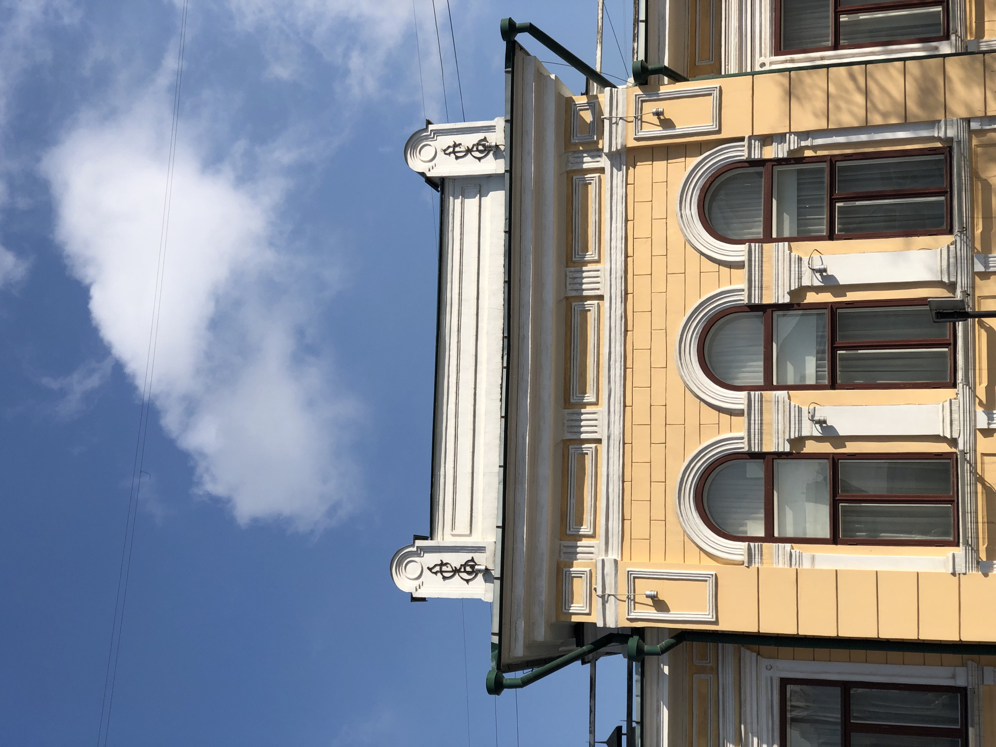 The image captures a detailed view of an ornate building facade against a bright blue sky with white, fluffy clouds. The building, located in Krasnoyarsk, Russia, is primarily light yellow, accented with elaborate white architectural trim, cornices, and decorative elements.
The facade features multiple windows, including several arched windows on the upper levels, each framed by white decorative surrounds. Adjacent to these are rectangular windows with dark brown frames, some appearing to have blinds or shutters. Below the arched windows, small light fixtures are mounted on the yellow walls within decorative white rectangular panels.
At the top of the building, a prominent white cornice with intricate designs extends horizontally, and a decorative element resembling stylized intertwined shapes or an ornate symbol is visible on a corner. Dark green drainpipes run along the building's edges and roofline.
The scene is bathed in bright daylight, casting distinct shadows on the architectural details, suggesting a clear, sunny day, likely midday or afternoon. Thin, dark utility wires are visible stretching across the blue sky in the upper left section of the frame. There is no visible text in the image.