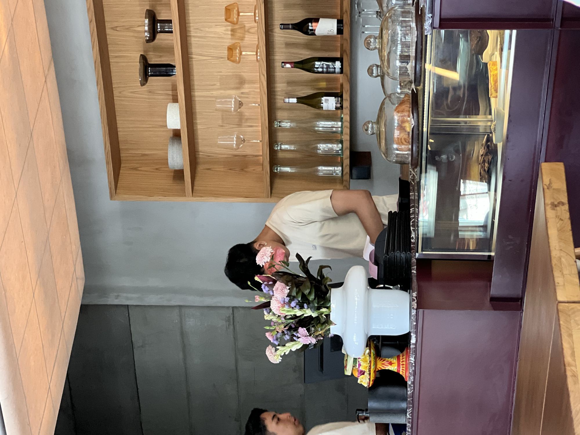 The image captures the interior of a modern cafe, likely located in North Kuta, Indonesia. In the foreground, a wooden table is visible. Behind it, a dark burgundy counter houses a glass display case filled with pastries and cakes. Two individuals are present: one, wearing a light-colored t-shirt, is standing behind the counter, appearing to be a barista or server, and is bent over, possibly organizing items or attending to a customer. Another person, also in a light-colored t-shirt, is partially visible on the left, looking towards the counter. A large white vase filled with a vibrant arrangement of pink and purple flowers sits prominently on the counter. Behind the counter, a wooden shelving unit displays various items, including glassware, bottles of wine, and decorative objects. The walls are a mix of textured gray concrete and a lighter gray finish. The ceiling is tiled with square panels, illuminated by warm lighting.